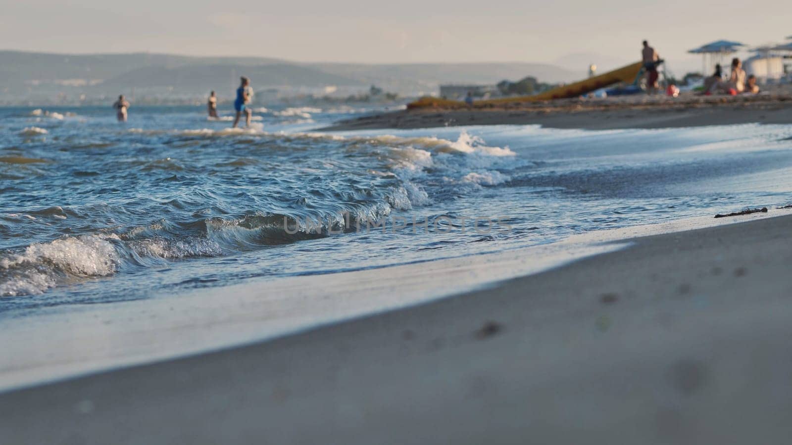 Small waves gently washing sandy beach of Crimea at sunset, with tourists enjoying summer vacation in the background