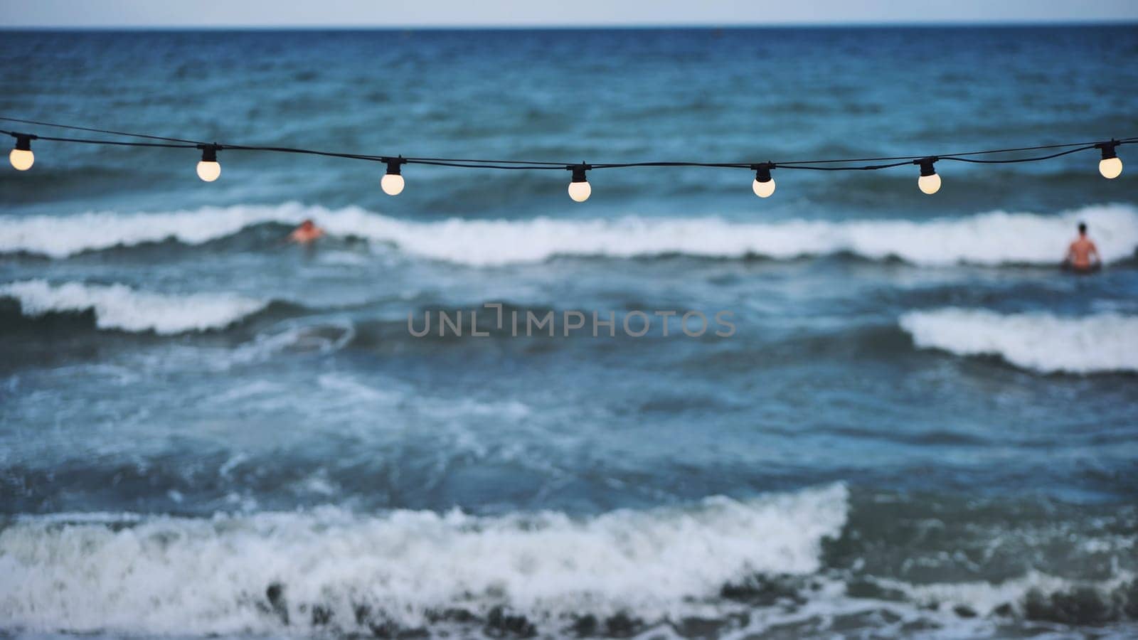 String of lights hanging over the crimean sea at dusk while tourists swim and enjoy the waves