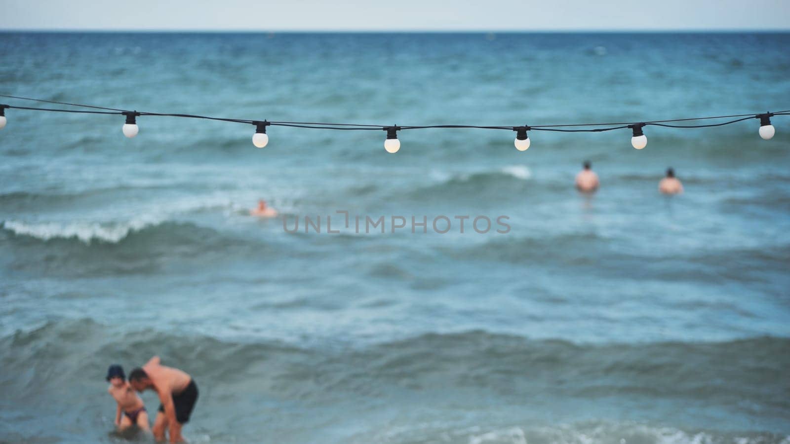 Father and son bathing in the sea in Crimea, Russia, with string lights and tourists in the background
