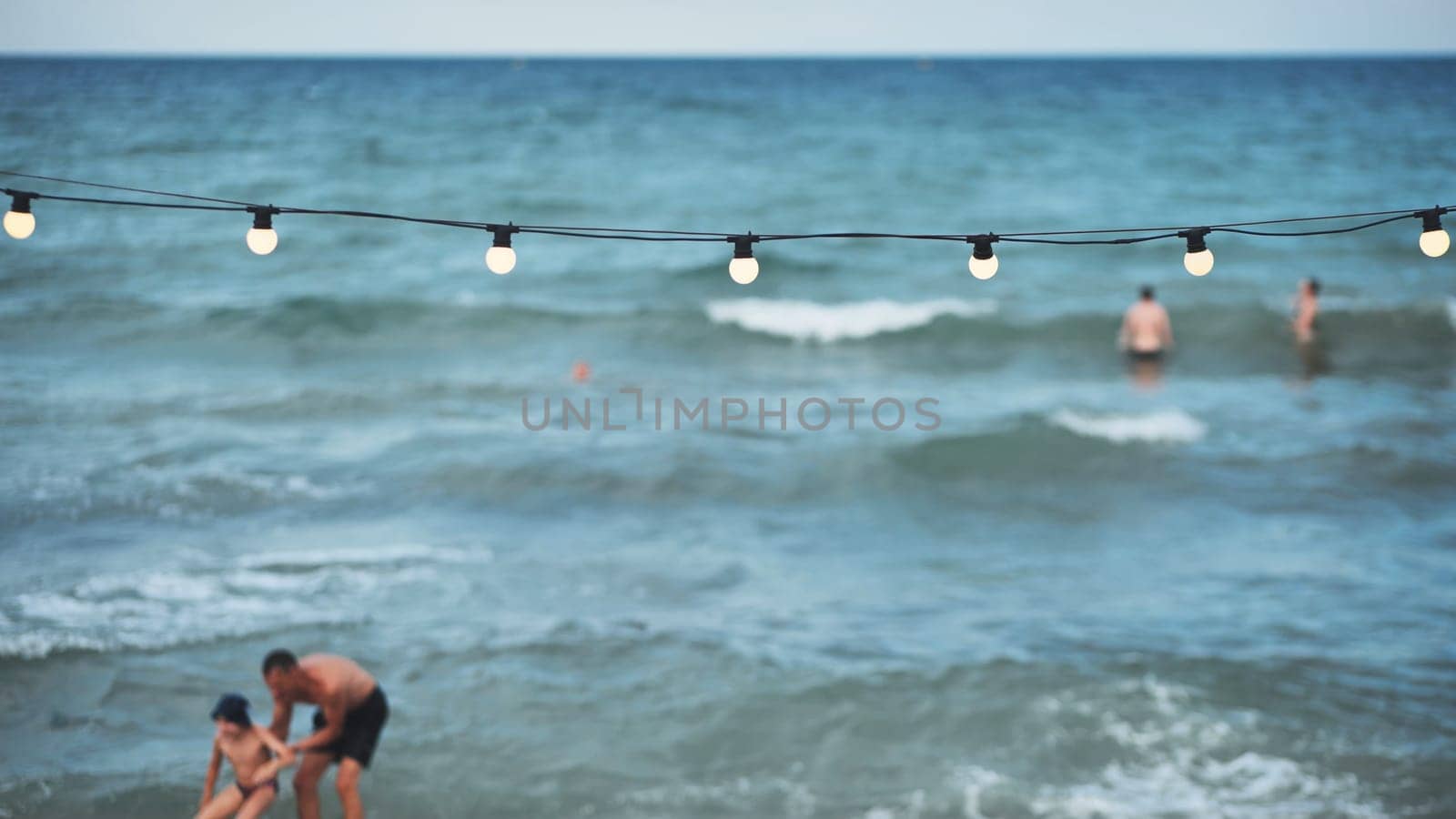 Father helping his son to stand up on the beach with string lights and blurred swimmers in the background in Crimea