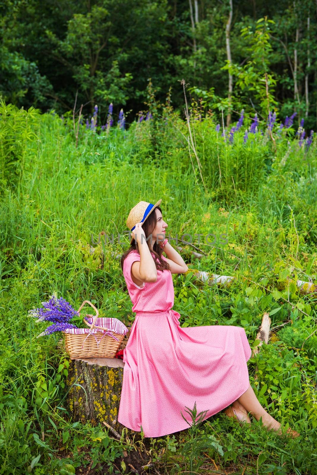A young woman in a pink dress sits on a tree stump in a sunny forest glade, enjoying a picnic with wildflowers.
