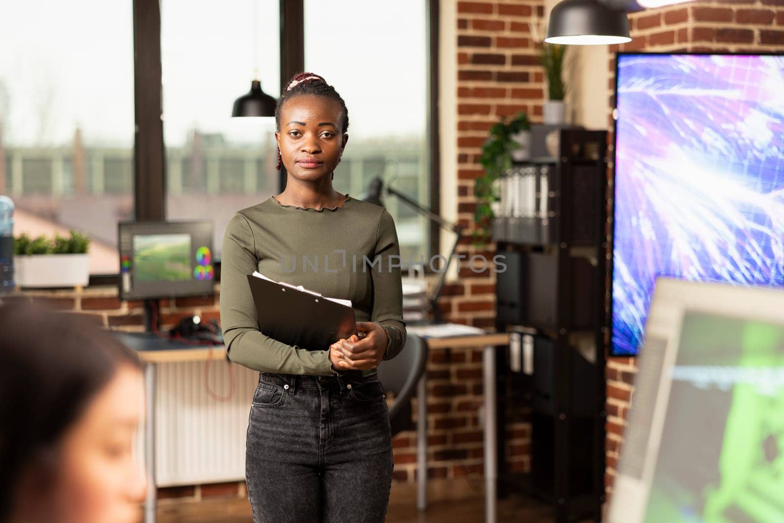 Portrait of female manager standing in modern office, holding a clipboard and posing as her coworker focuses on their computer. Black professional woman in brick wall workspace, looking at camera.