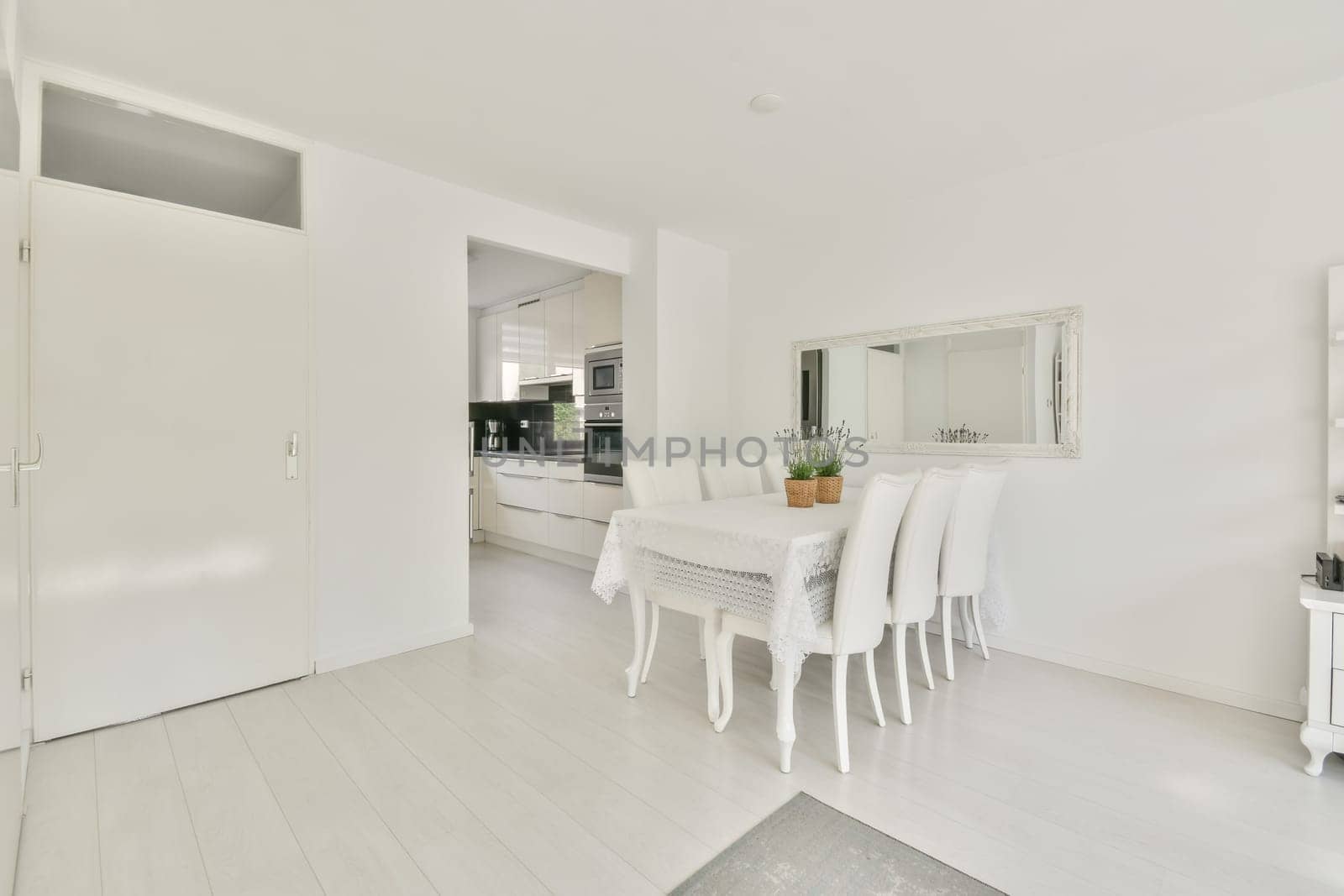 a white dining room with wooden flooring and large mirror on the wall above it is an open kitchen area