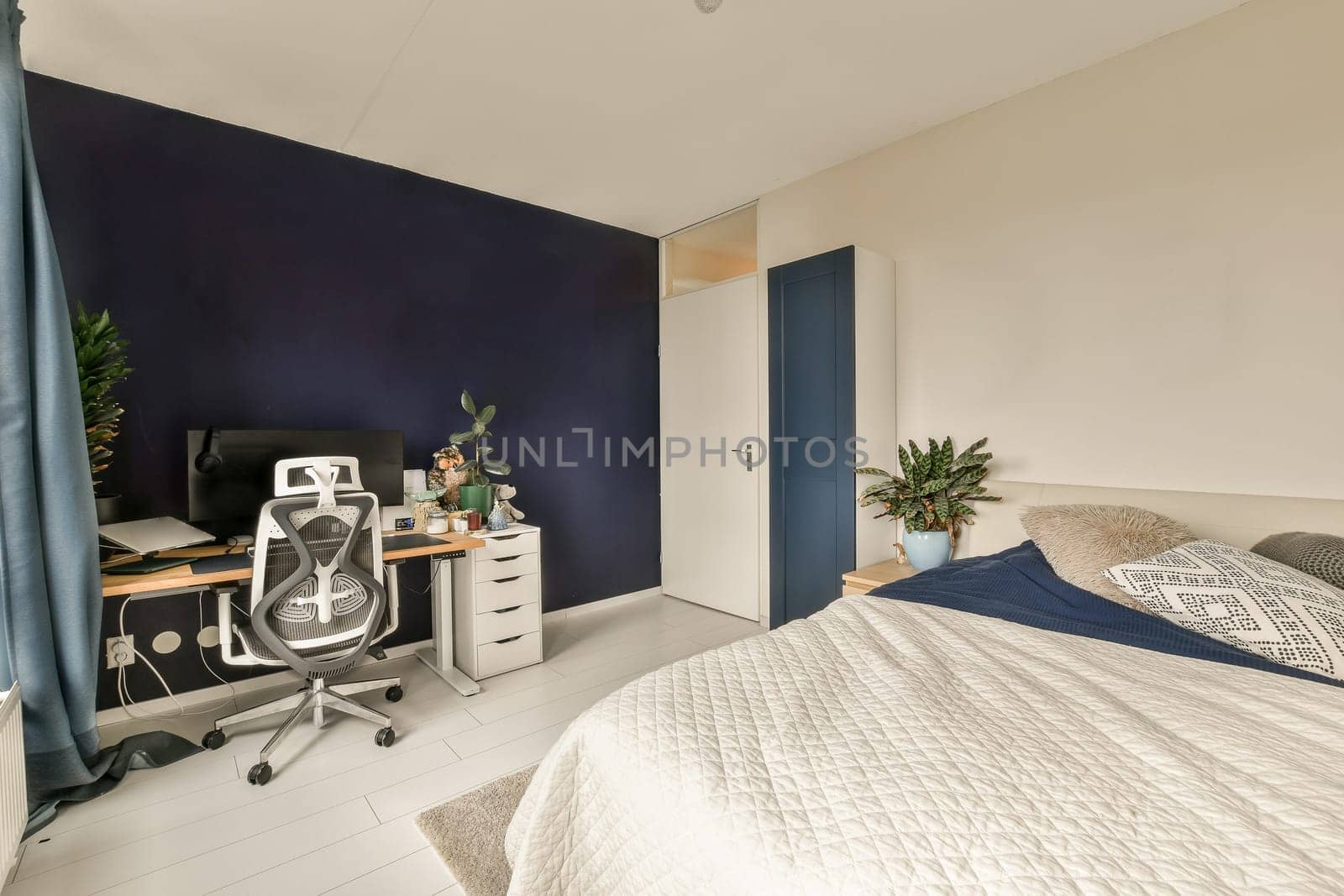 a bedroom with a bed, desk and computer monitor on the wall in front of the bed is dark blue