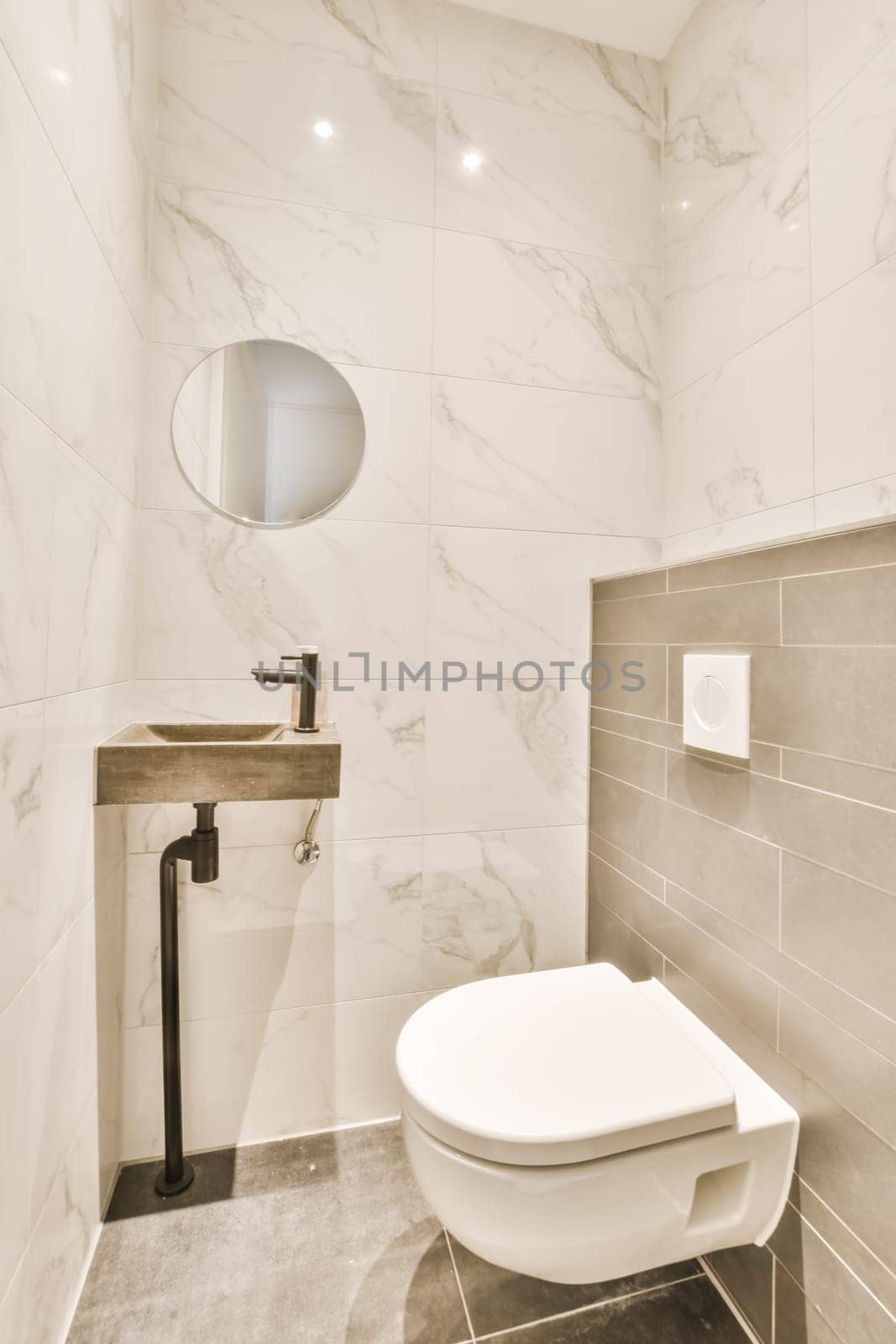 a white toilet in a small bathroom with marble tiles on the walls, and a round mirror hanging above it