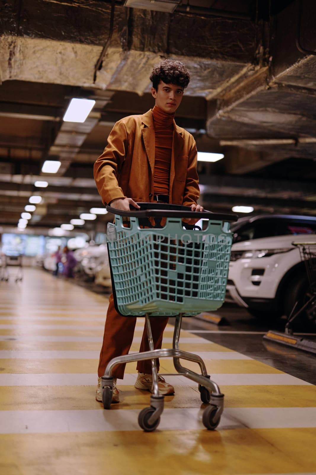 A fashionable male model poses with a shopping cart in a modern parking garage The setting includes parked cars and vibrant yellow and gray lines on the floor