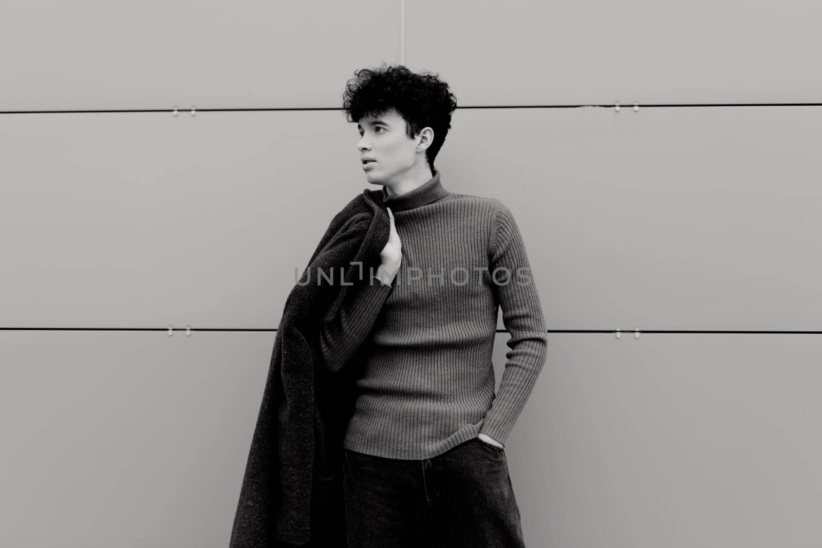 Fashion photo of a young man in black and white, leaning against a wall with hands in pockets and jacket over shoulders.