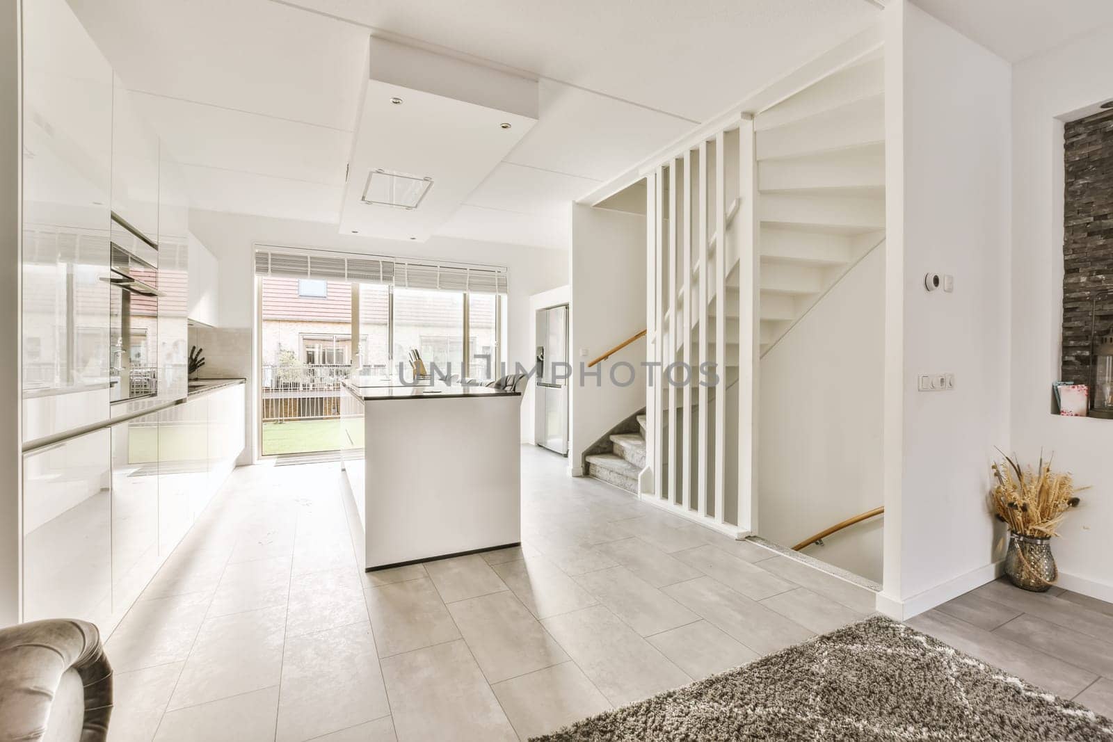 a living room with white walls and grey flooring in the center of the room is an open kitchen area