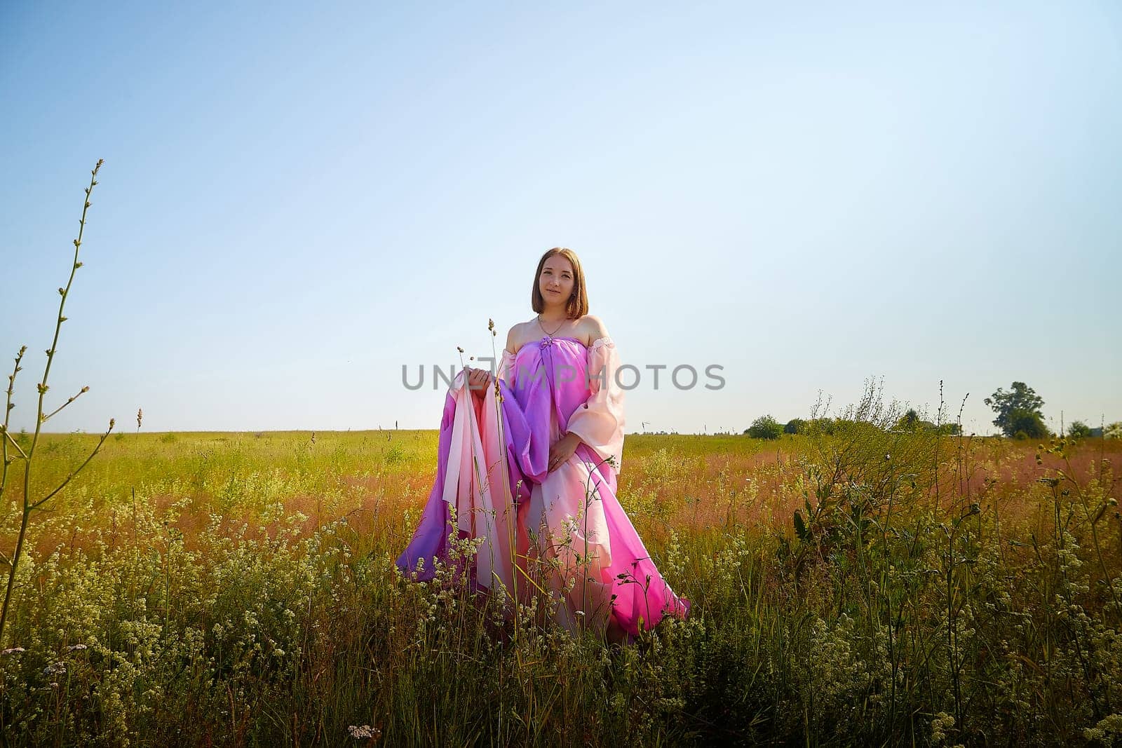 A woman wearing a colorful flowing garment enjoys the warm sunlight in a lush green field, surrounded by wildflowers and clear skies.