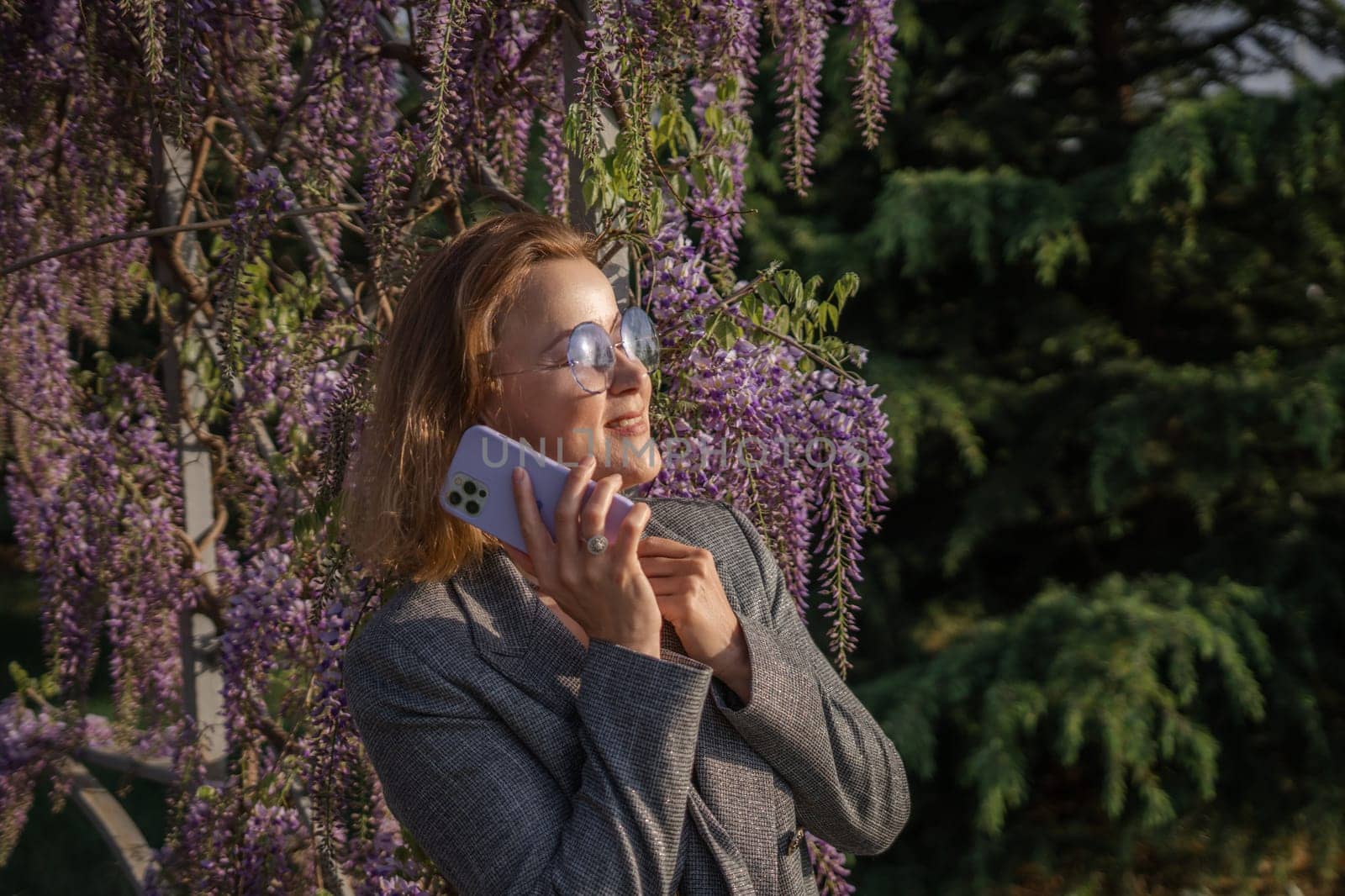 A woman in sunglasses and a blazer stands in front of a wall of purple wisteria, holding a white phone to her ear. She is looking up and smiling.