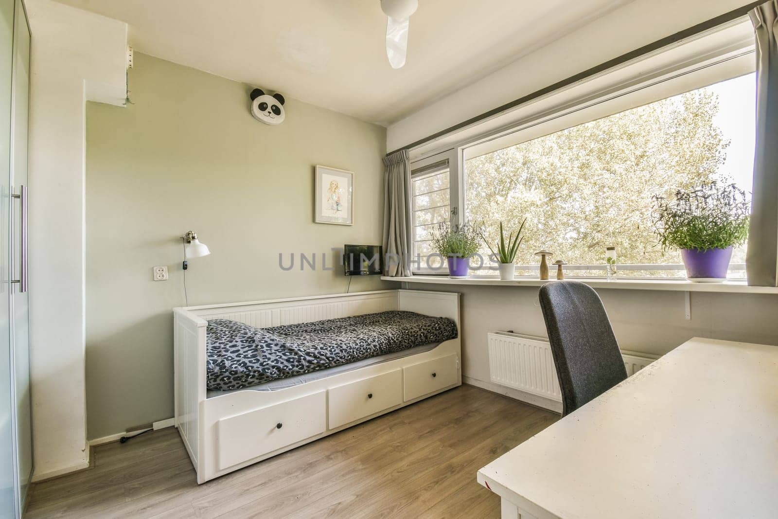 a small room with a desk and some plants in the window simh is on the wall next to the bed
