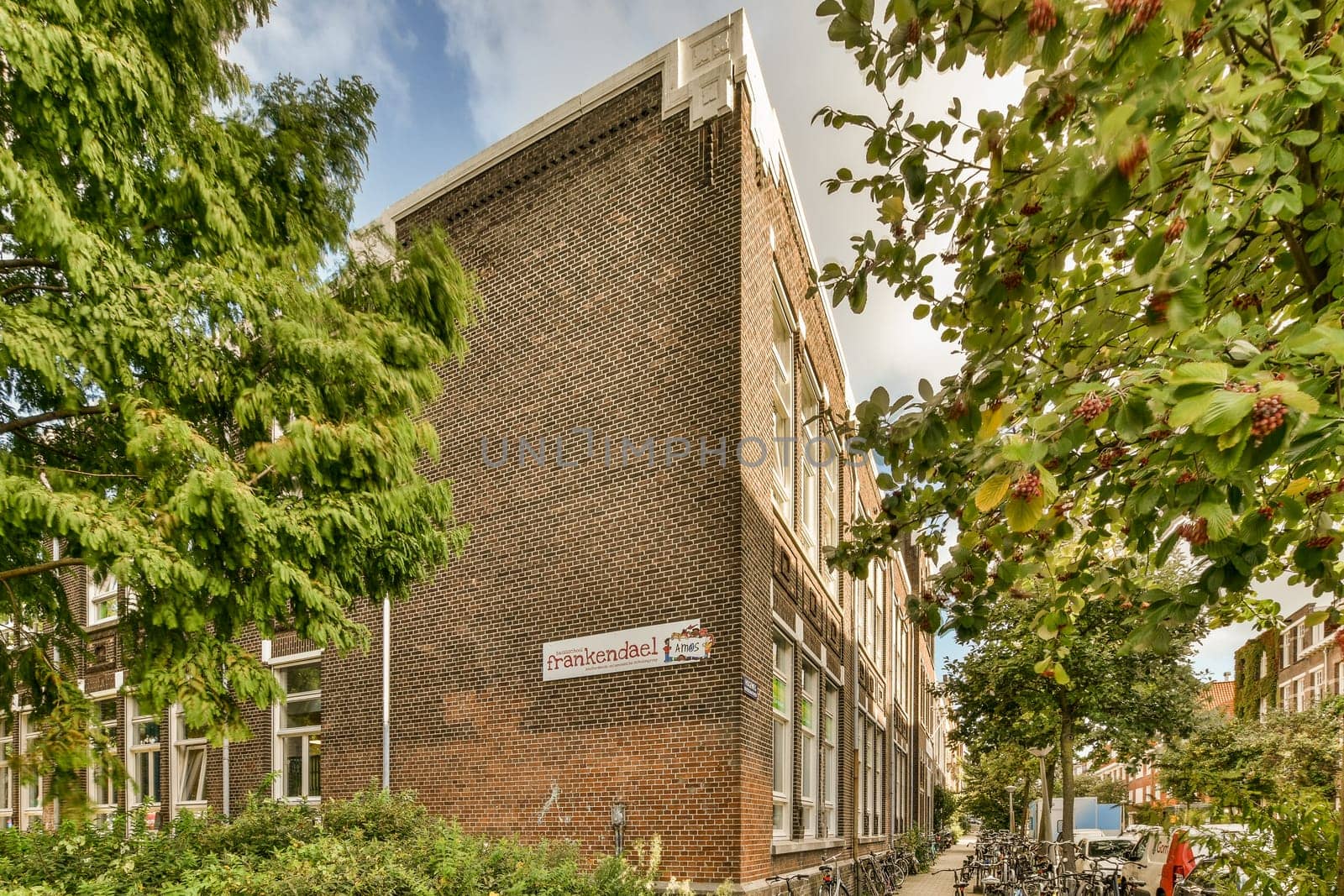 a brick building with bicycles parked on the sidewalk next to trees and bushes in front of it, under a cloudy blue sky