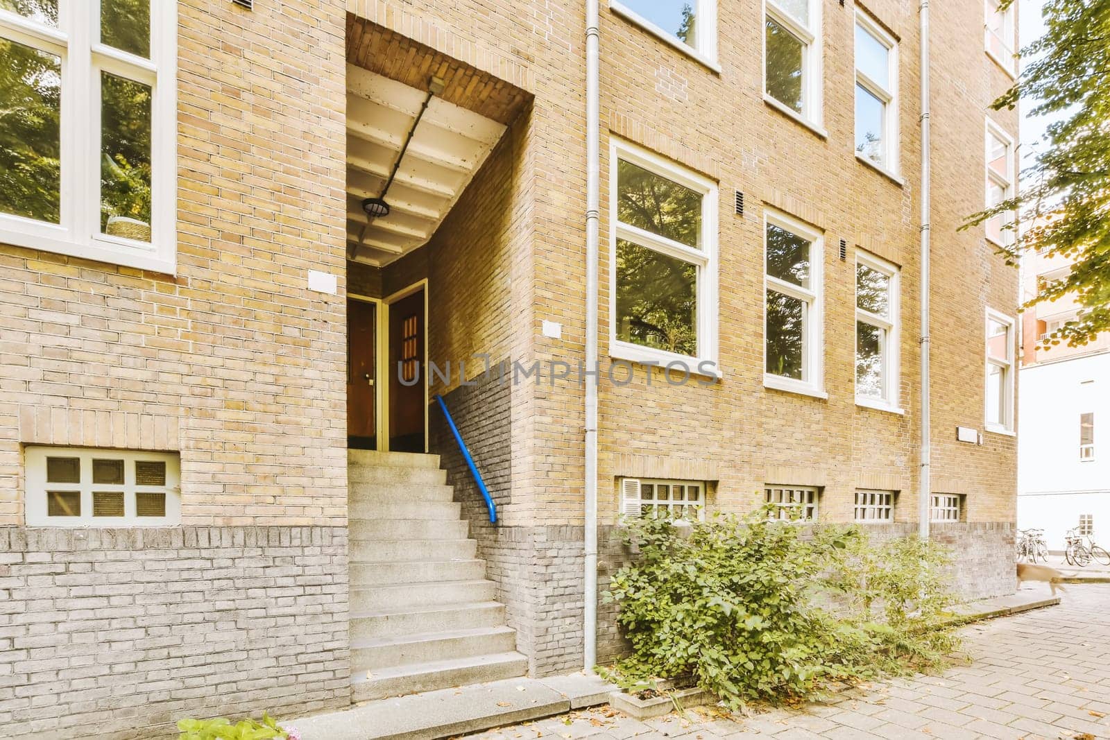 a brick building with some plants growing in the front and stairs leading up to the second floor, on a sunny day