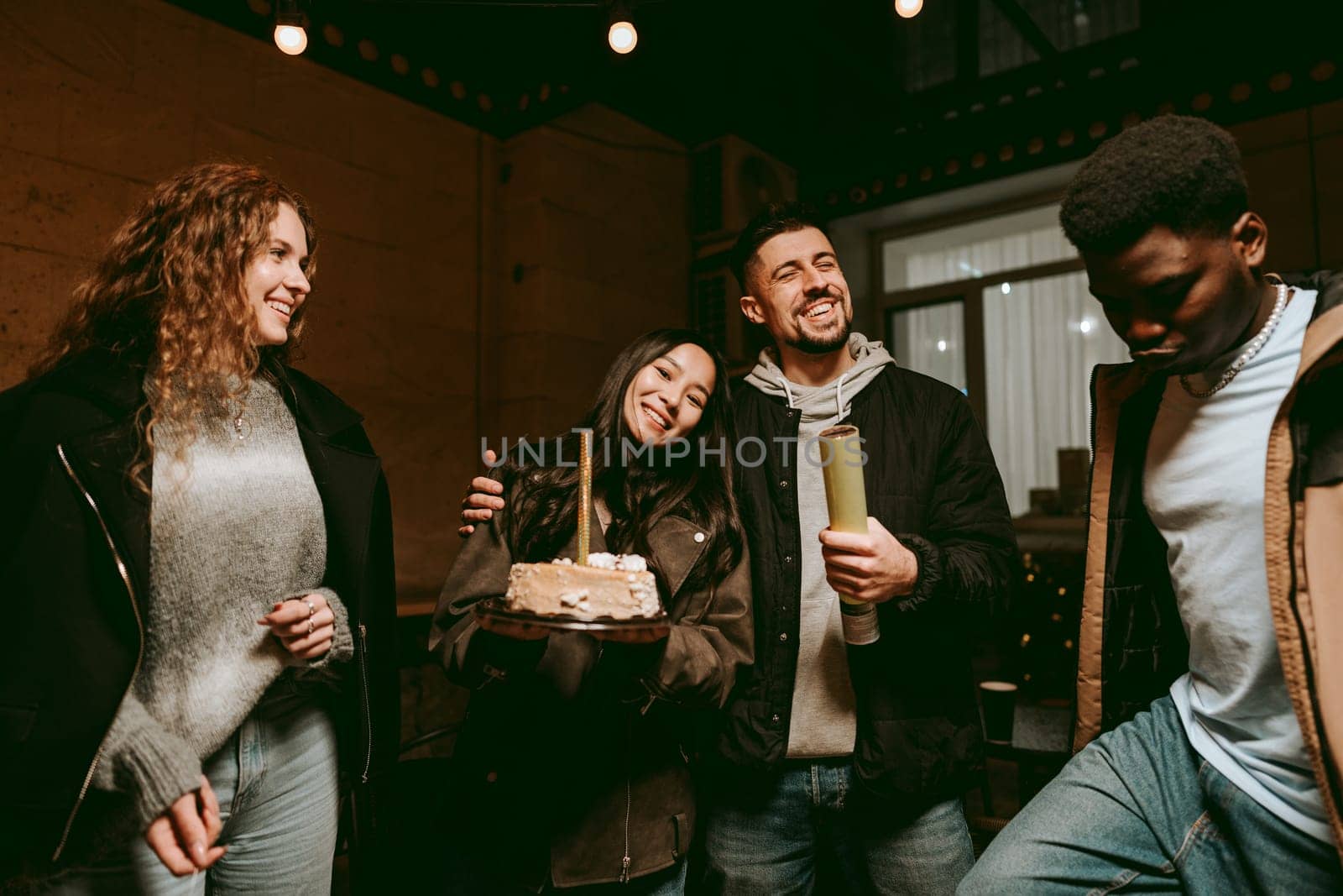 Group of four friends enjoy a cozy evening indoors, celebrating with a cake. They share laughter and joy as they pose together in warm, inviting light.