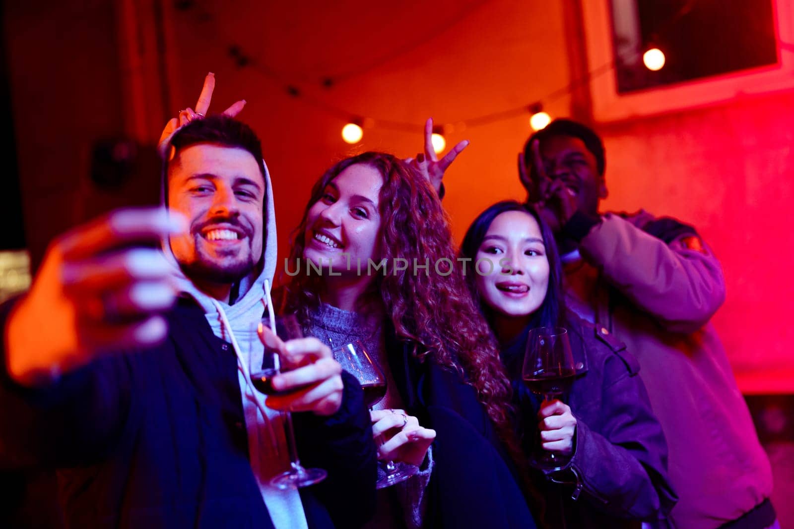 Group of friends celebrating at a lively party in an urban area, holding wine glasses and posing cheerfully under colorful lights in the evening.