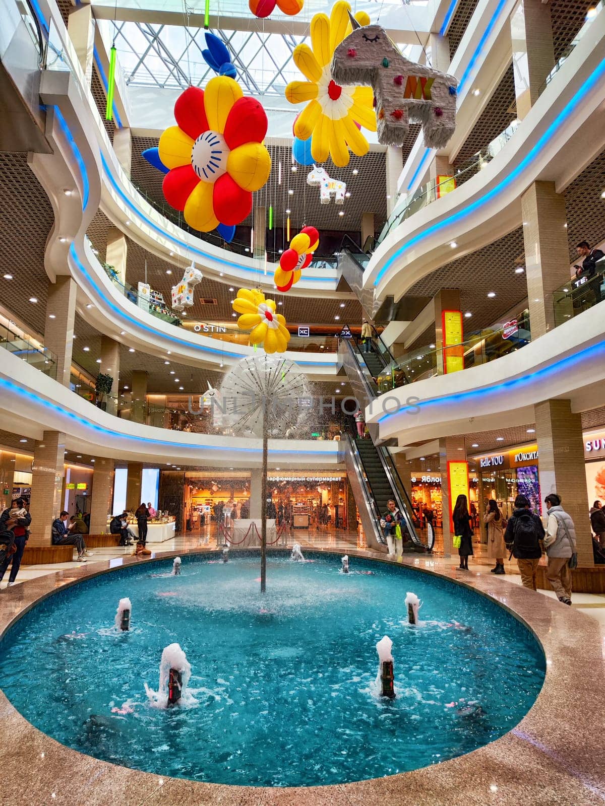 Irkutsk, Russia - April 04, 2025: Visitors explore a lively shopping center in Irkutsk, featuring a central fountain and colorful decorations hanging from the ceiling.