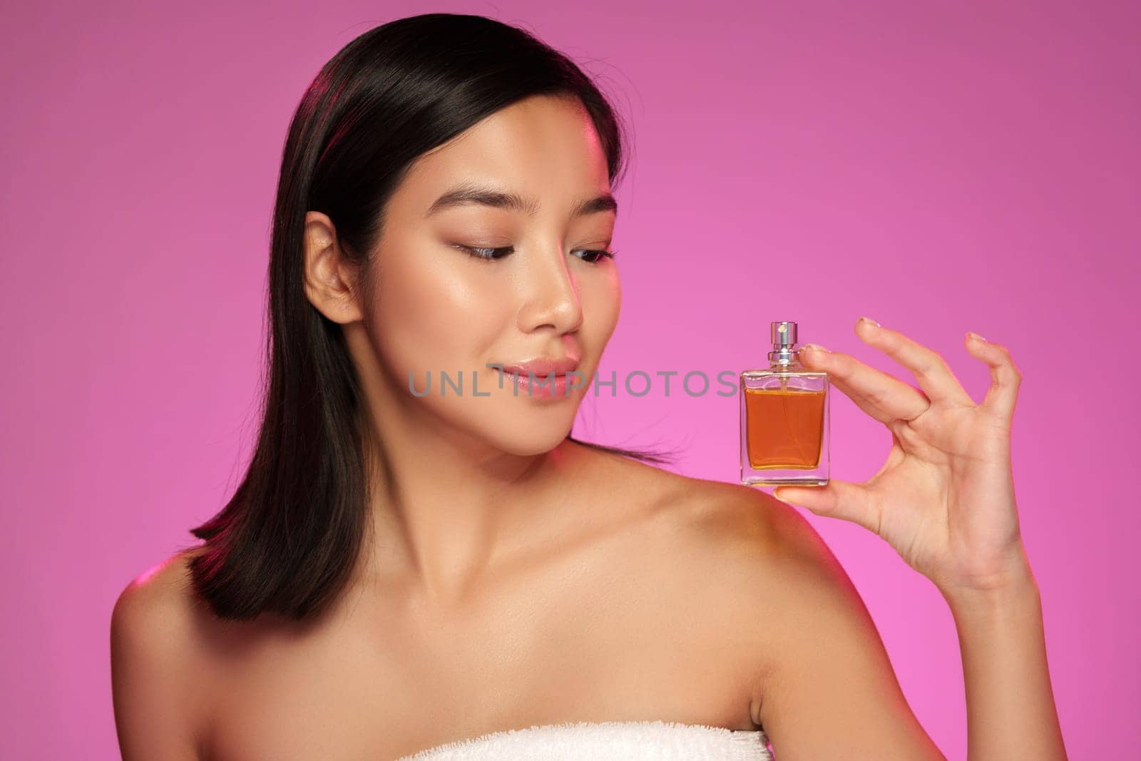 A young woman is showcasing a perfume bottle against a bright pink backdrop. She appears confident while gently holding the bottle, highlighting its elegance.