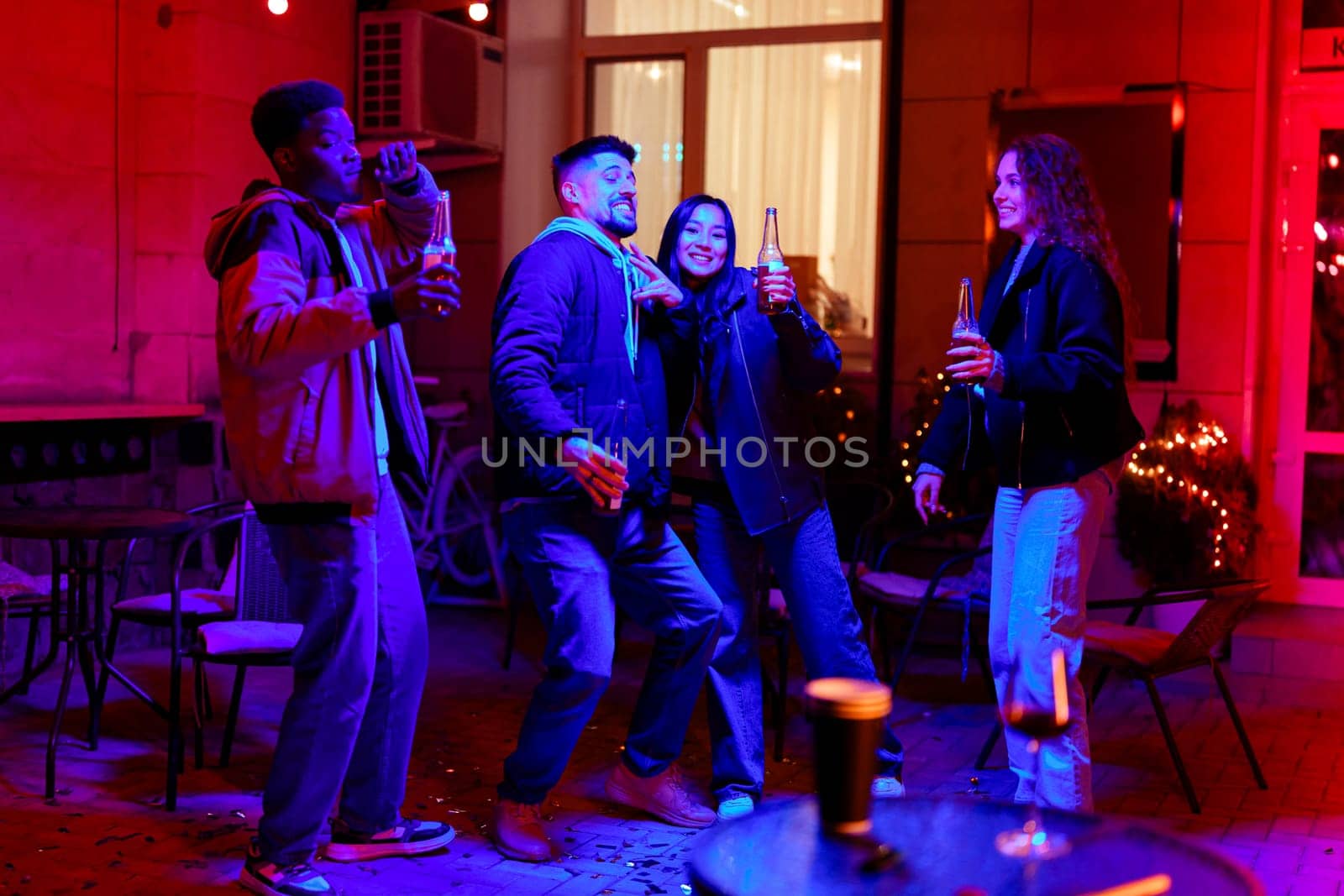 Four friends enjoy a lively evening gathering filled with laughter and drinks. The colorful ambiance adds to the festive atmosphere as they celebrate together outdoors.