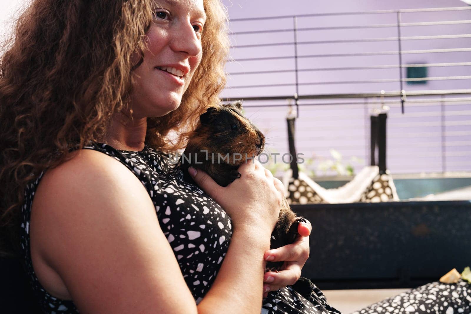A young girl smiles as she gently cradles a guinea pig close to her chest. She sits comfortably beside a pet cage, creating a warm atmosphere for bonding with her furry friend.