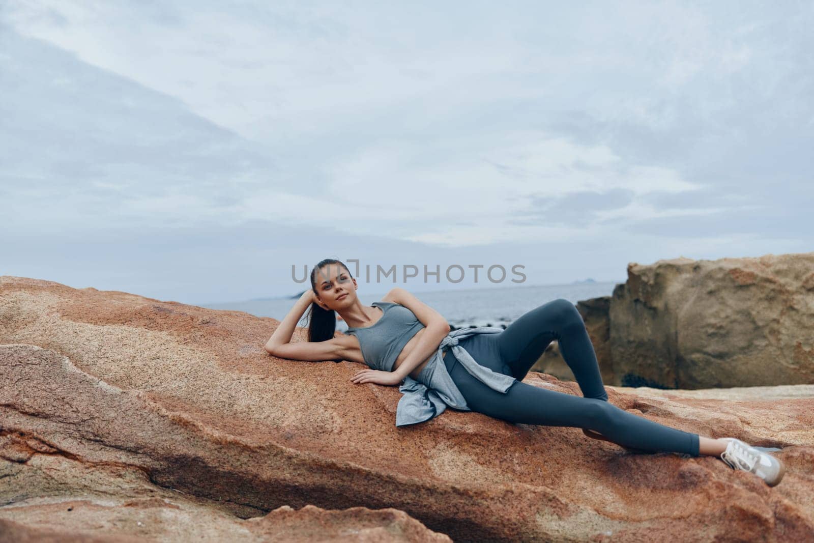 Fit female model in stylish sportswear lounges on coastal rocks, evoking relaxation and a healthy lifestyle against a tranquil ocean backdrop