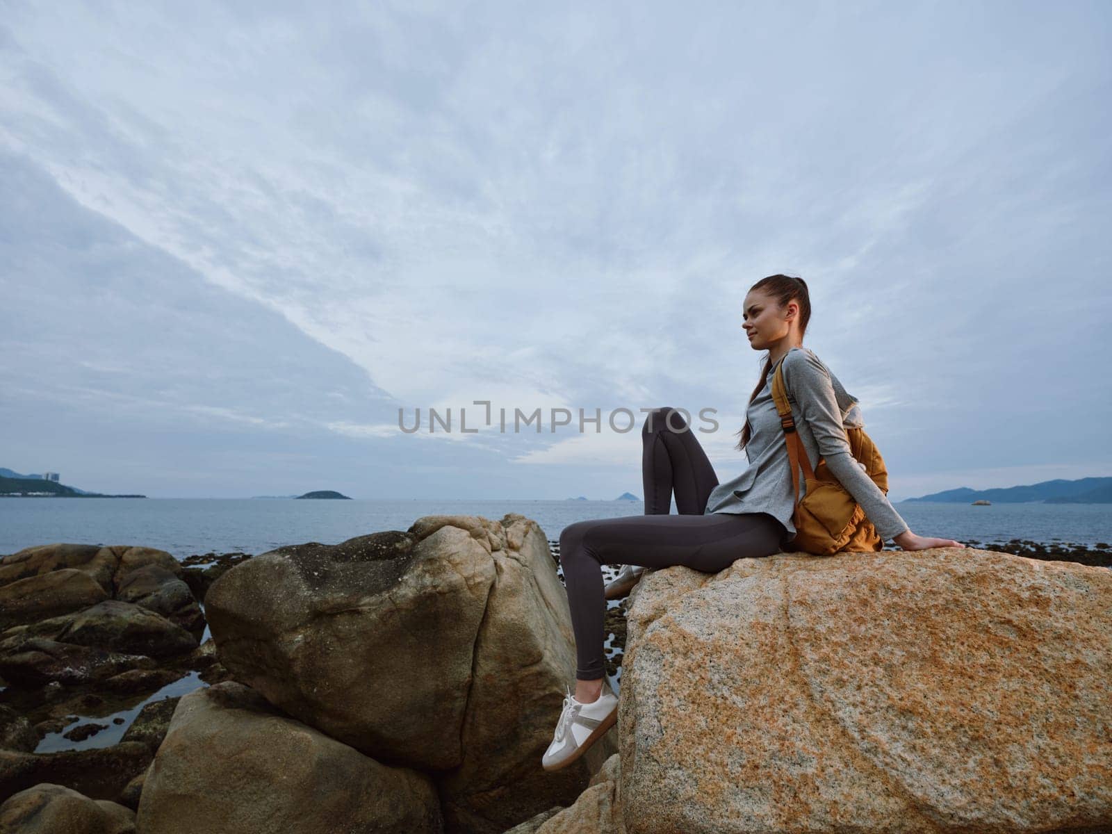 A young woman in casual outdoor clothing sits on a rock by the sea, gazing at the horizon The scene evokes a sense of peace, adventure, and connection with nature