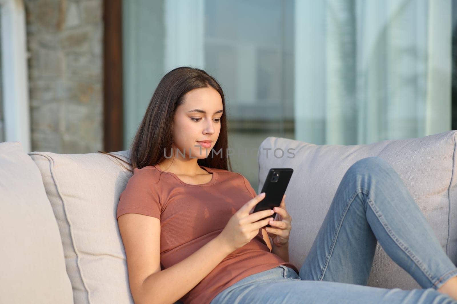 Relaxed woman in a balcony checking mobile phone sitting on a couch at home