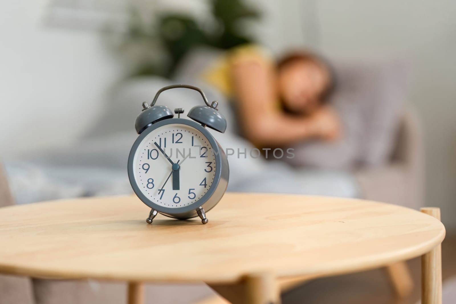 A classic alarm clock sits prominently, marking the beginning of a morning routine. The blurred figure of a woman in the background hints at her waking up.