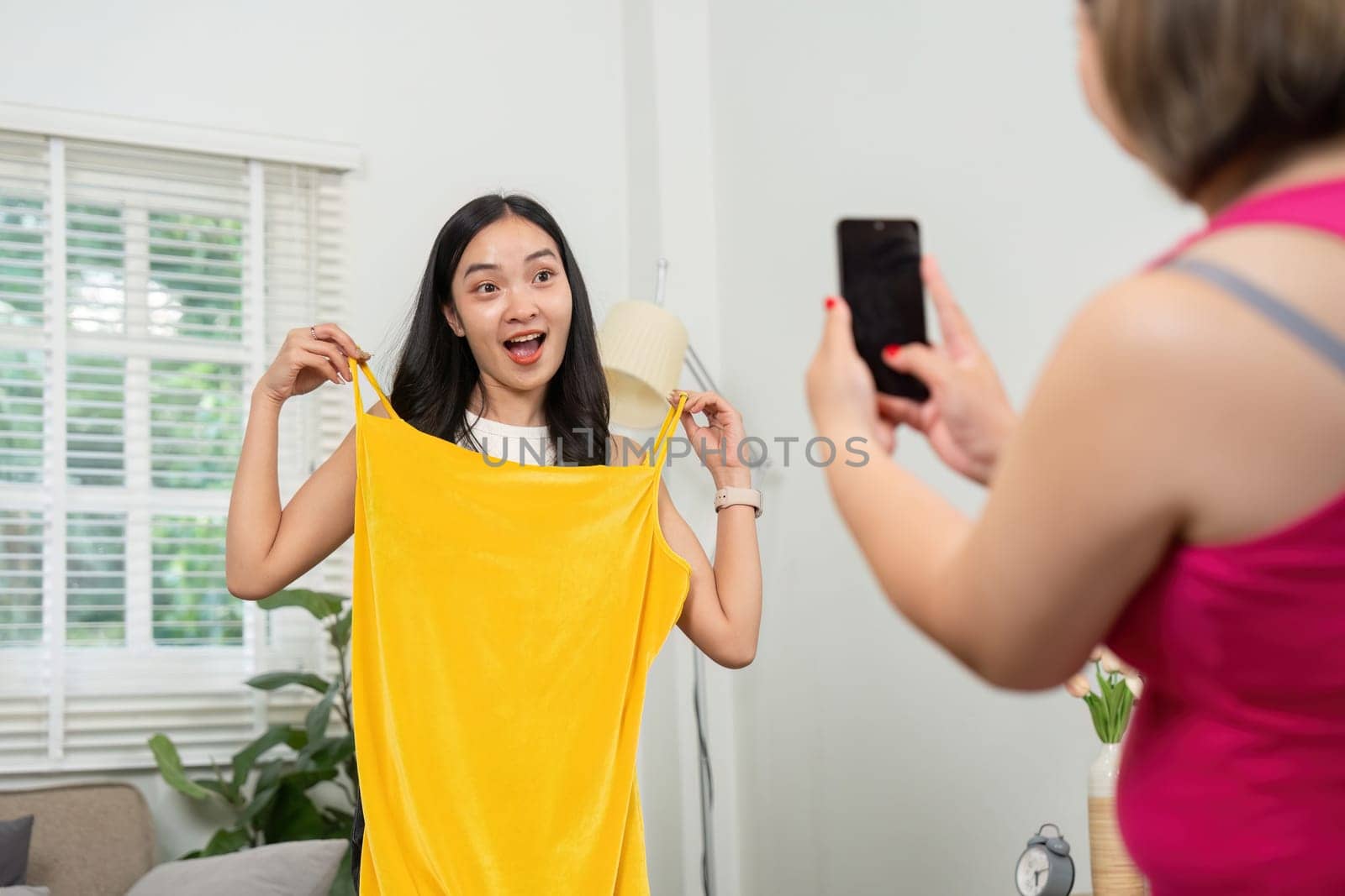 In a stylish living room, a woman joyfully holds up a vibrant yellow dress, engaging with her friend who is taking a photo. This moment captures the joy of fashion and friendship.