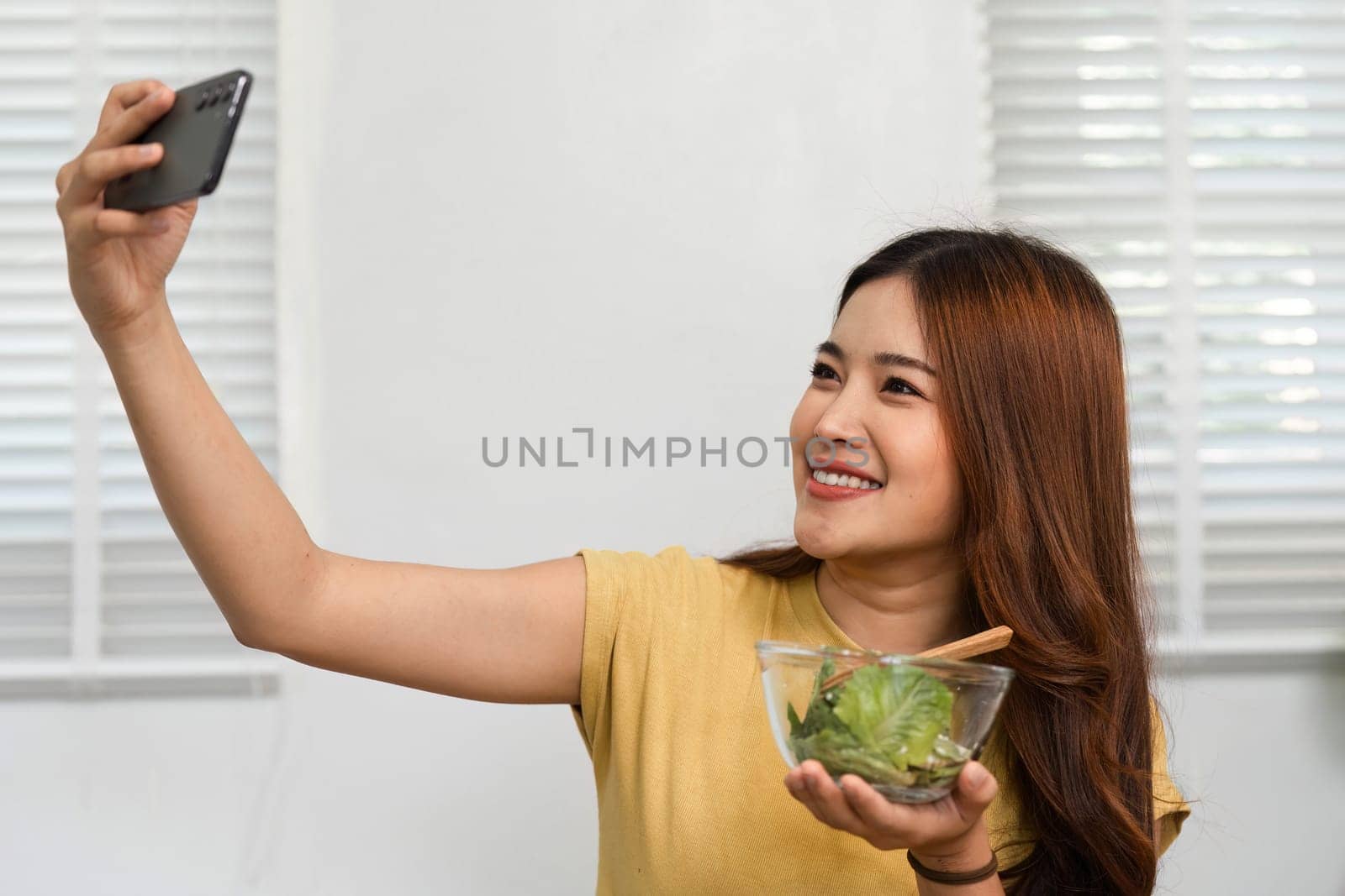 A joyful woman takes a selfie while savoring her nutritious salad, highlighting the blend of health and social media in her morning ritual.