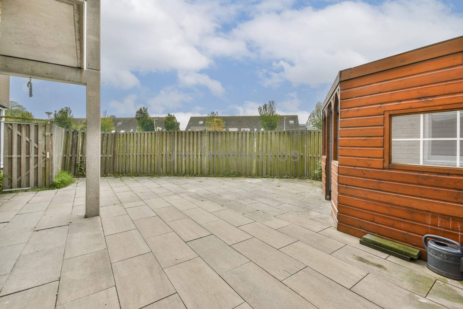 A tranquil outdoor area featuring a wooden structure, paved flooring, and a backdrop of greenery under a blue sky. Perfect for relaxation or gatherings.