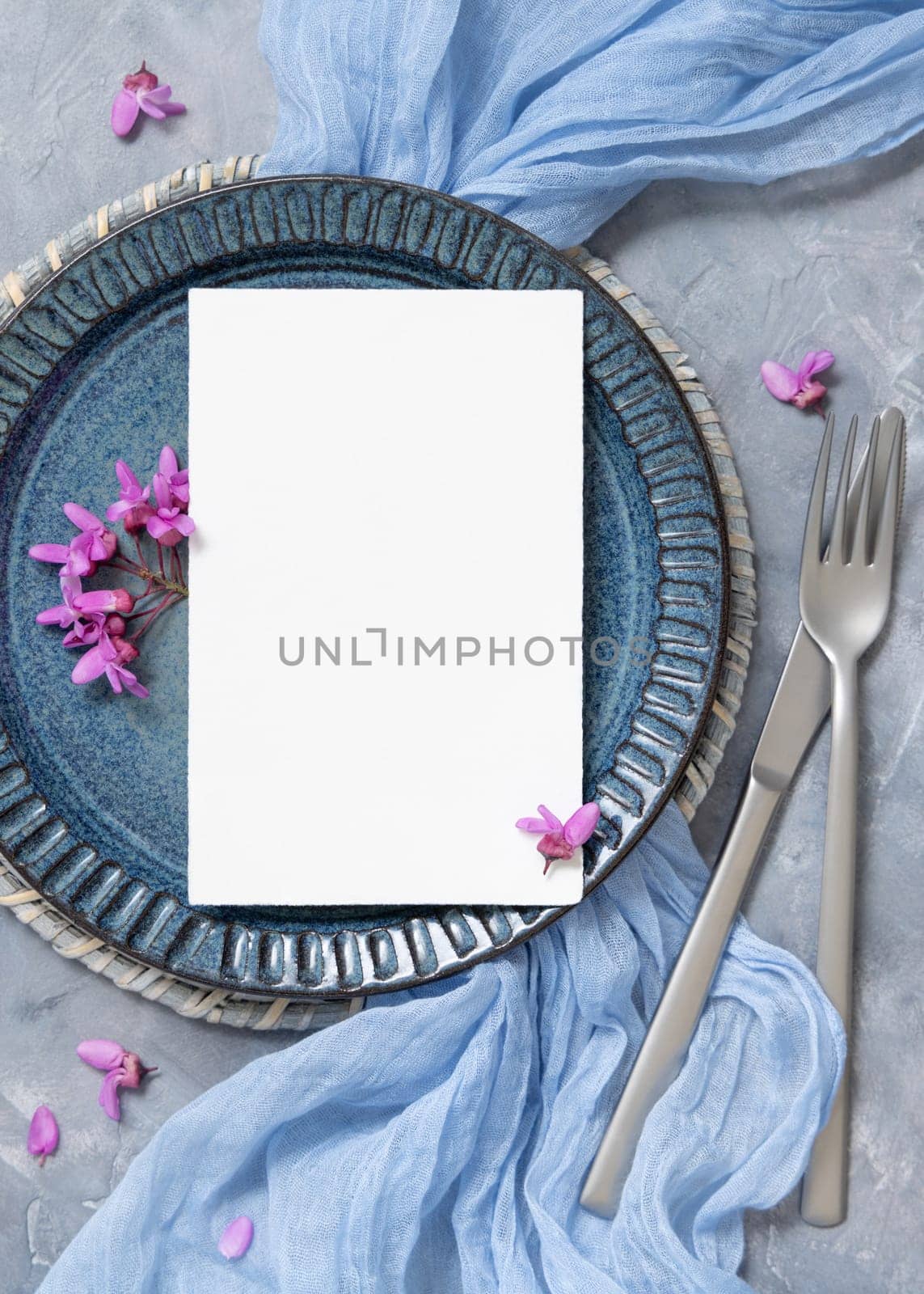 Pastel wedding table place with a card decorated with light blue fabric and wild purple flowers top view. Vertical blank card mockup for spring party design or romantic wedding stationery