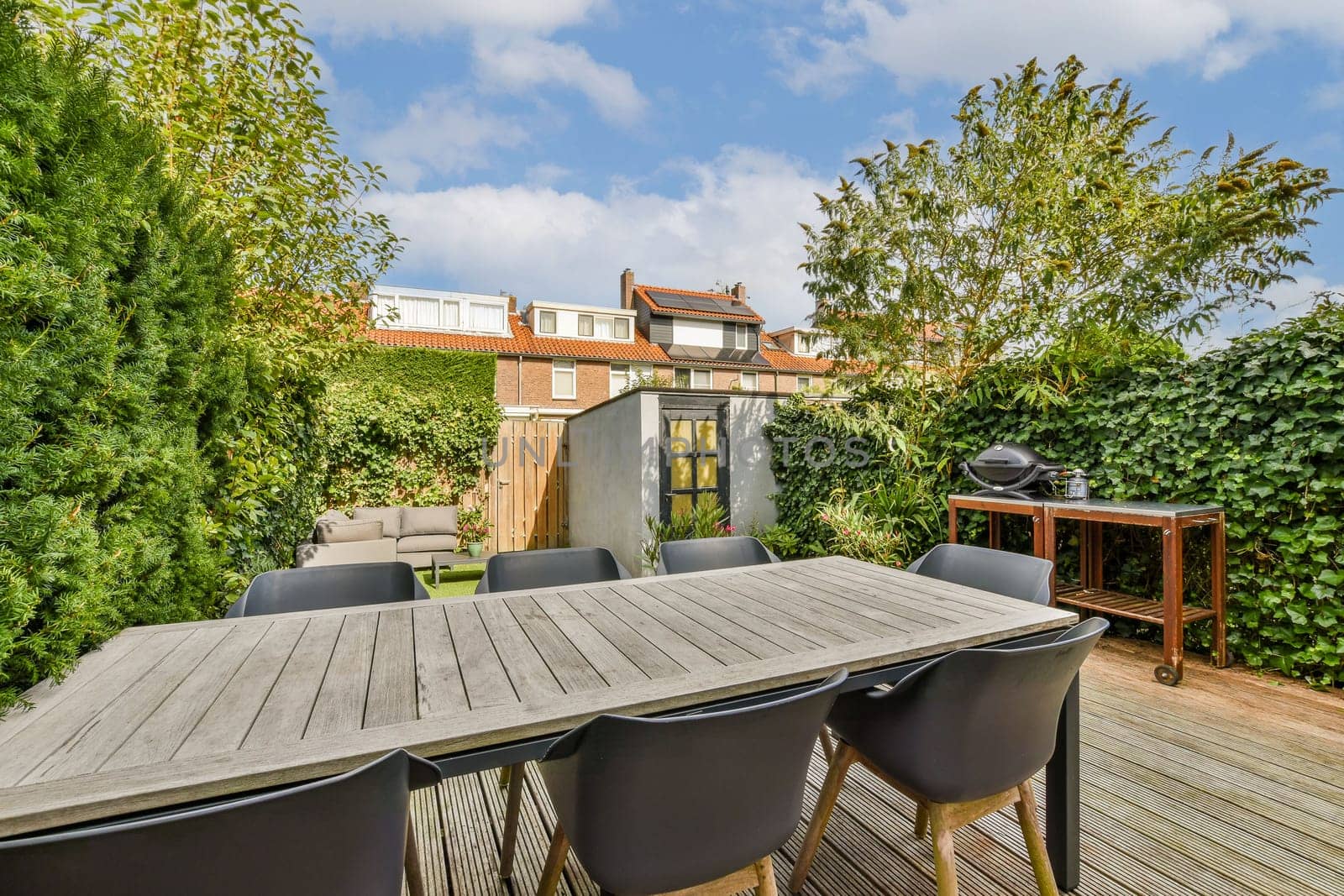 A stylish outdoor patio featuring a spacious wooden table surrounded by black chairs, set among lush greenery and blue skies. Ideal for gatherings and relaxation.