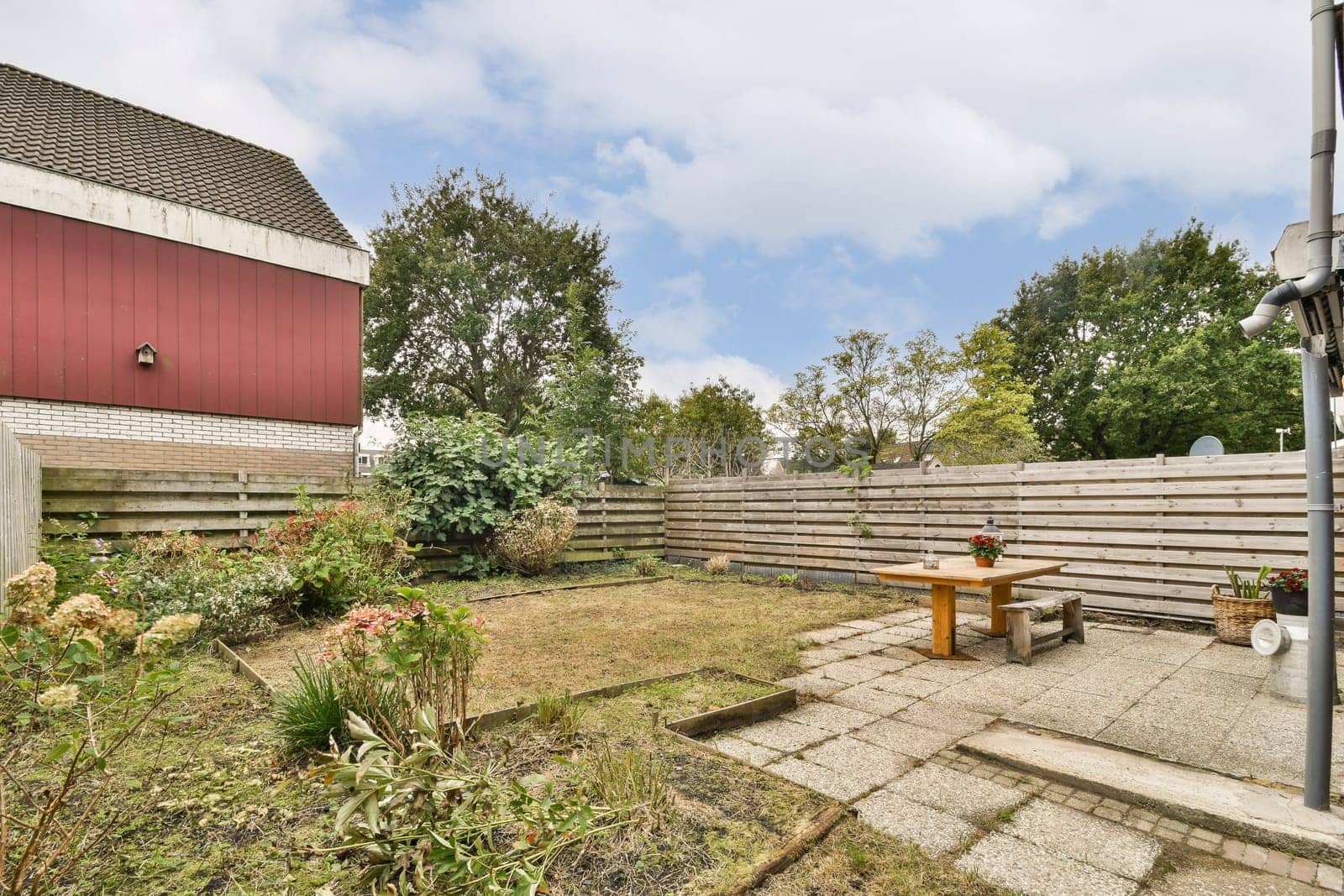 A tranquil garden scene featuring a wooden table and various decorative plants amidst a scenic backdrop of trees and a red building.