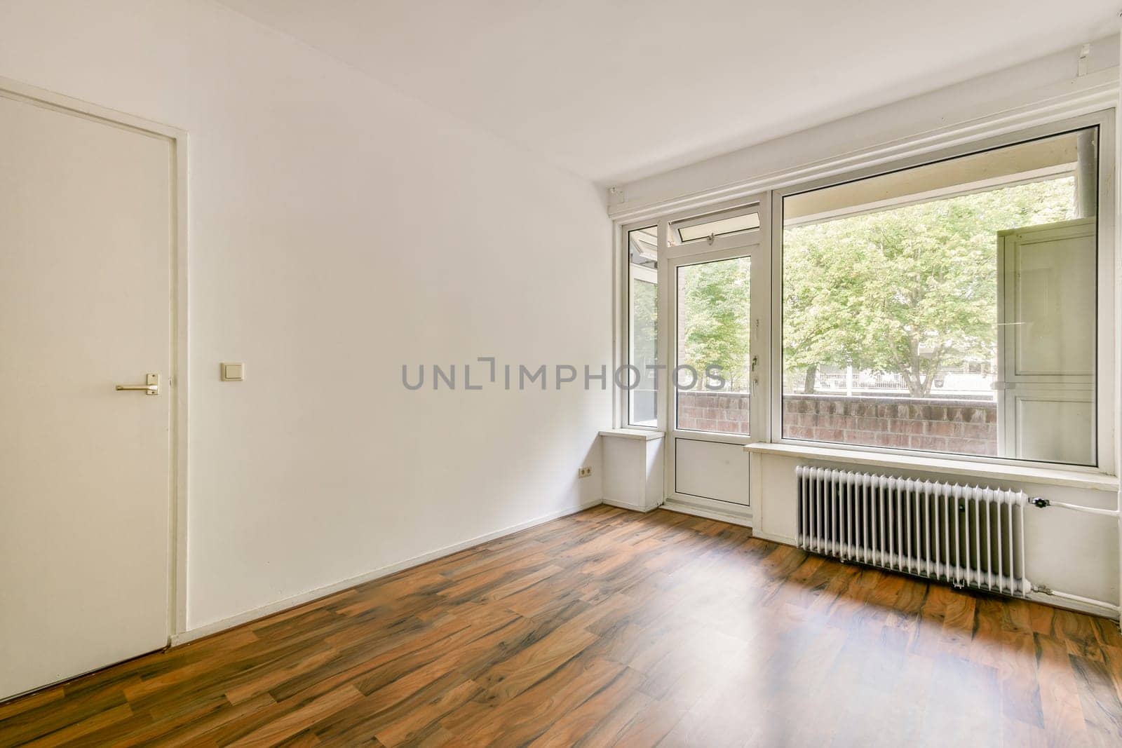 A minimalist interior featuring a large window allowing natural light to flood the room. The wooden floor adds warmth to the space, enhancing its inviting atmosphere.
