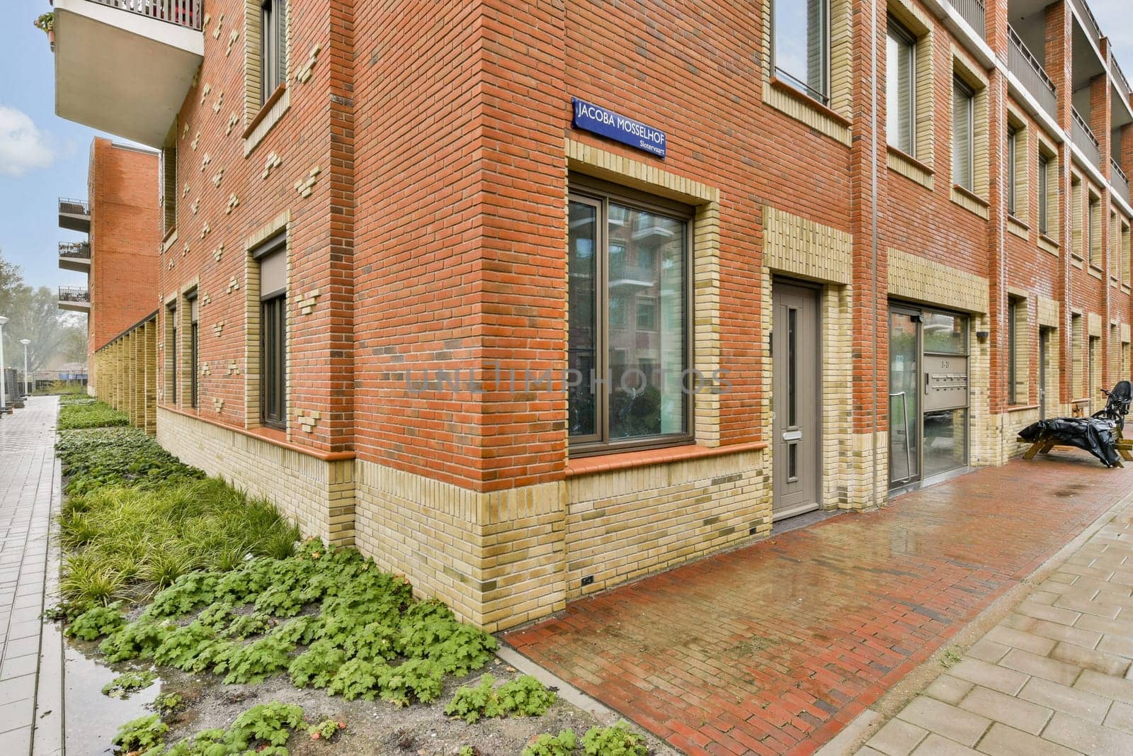 This illustration showcases a contemporary red brick building facade with large windows and greenery surrounding the entrance, emphasizing architectural design.