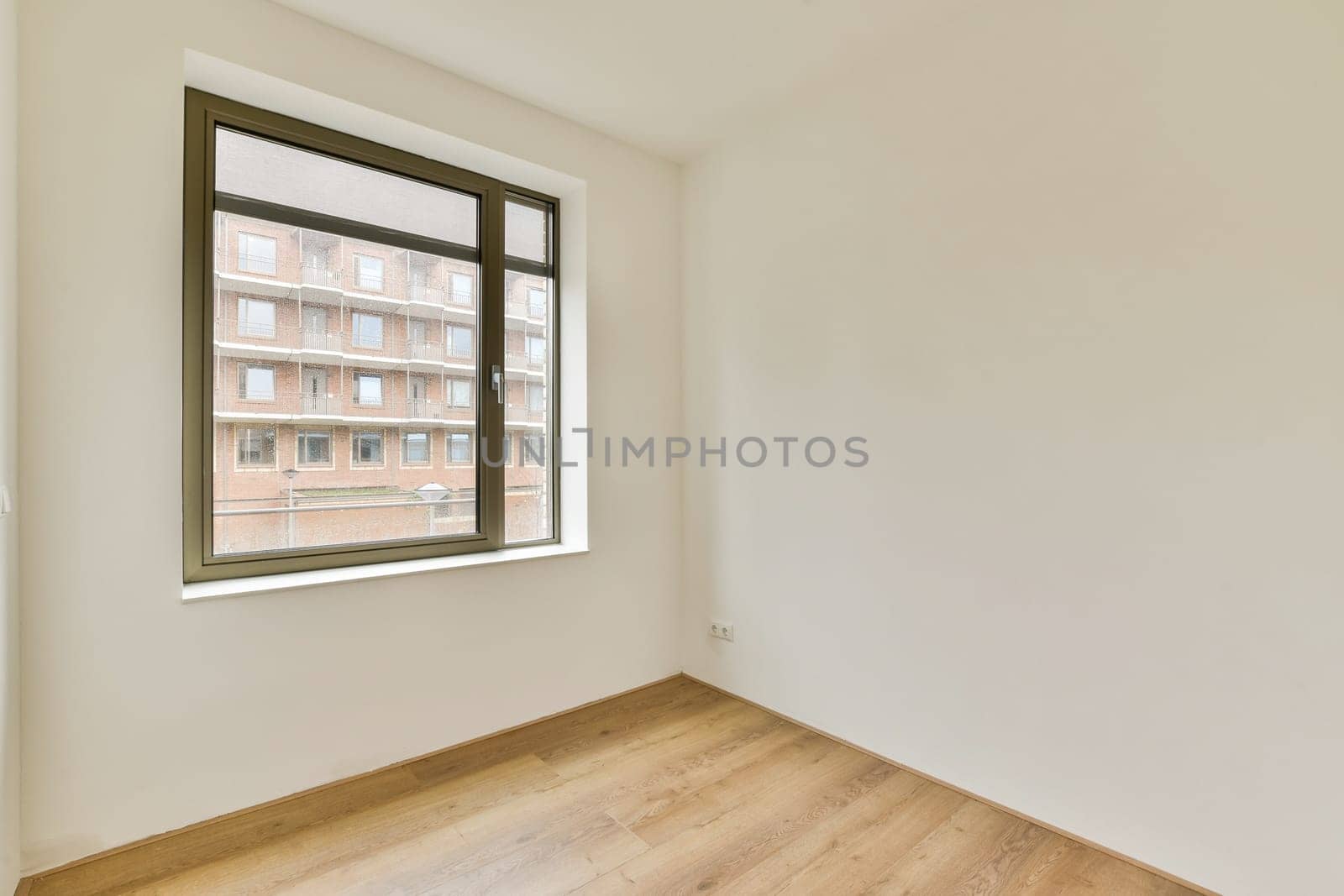 A spacious, minimally decorated room featuring a large window that lets in natural light, creating a bright atmosphere with wooden flooring.