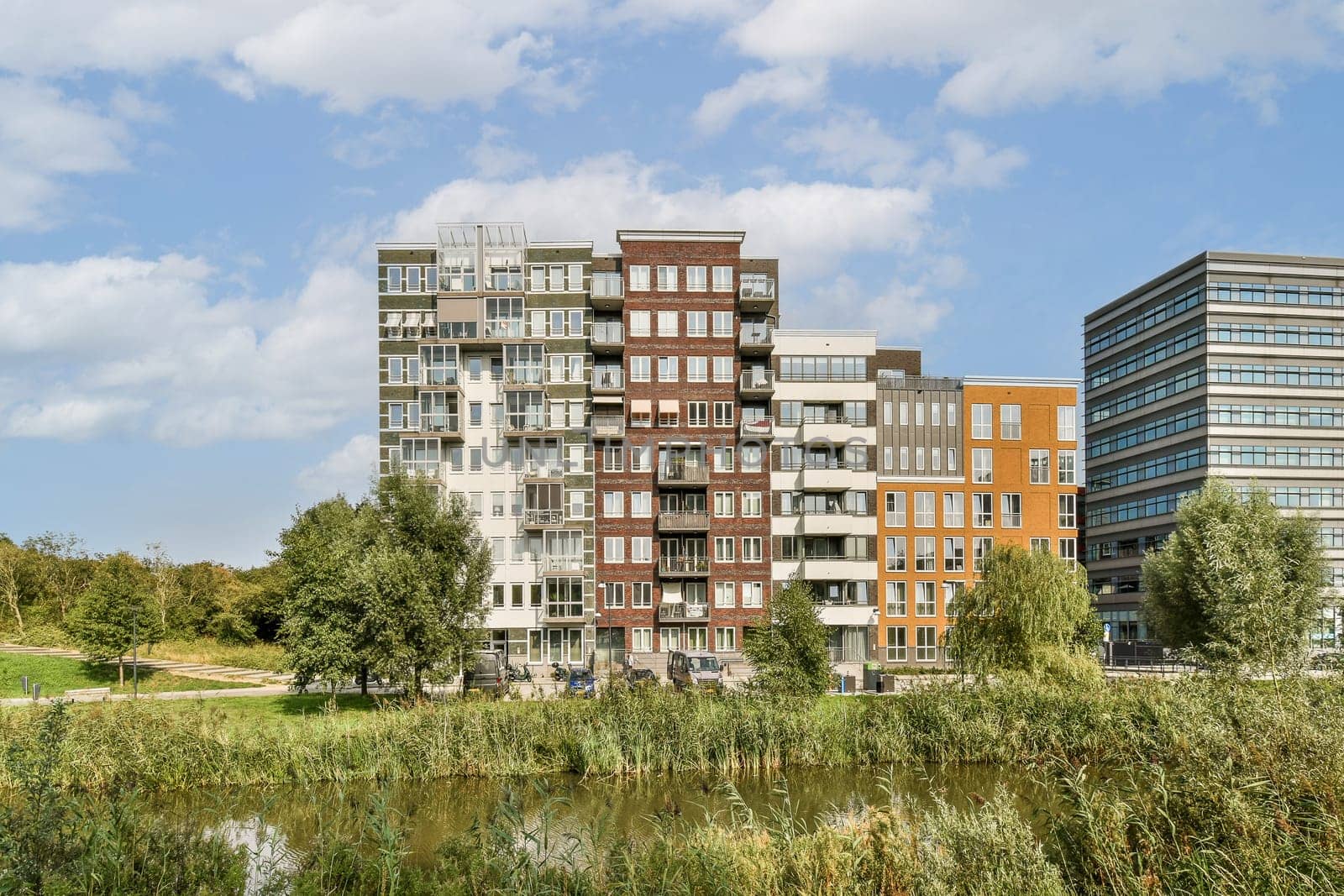 Modern apartment buildings stand by a serene park, showcasing architectural diversity and green space integration near water.