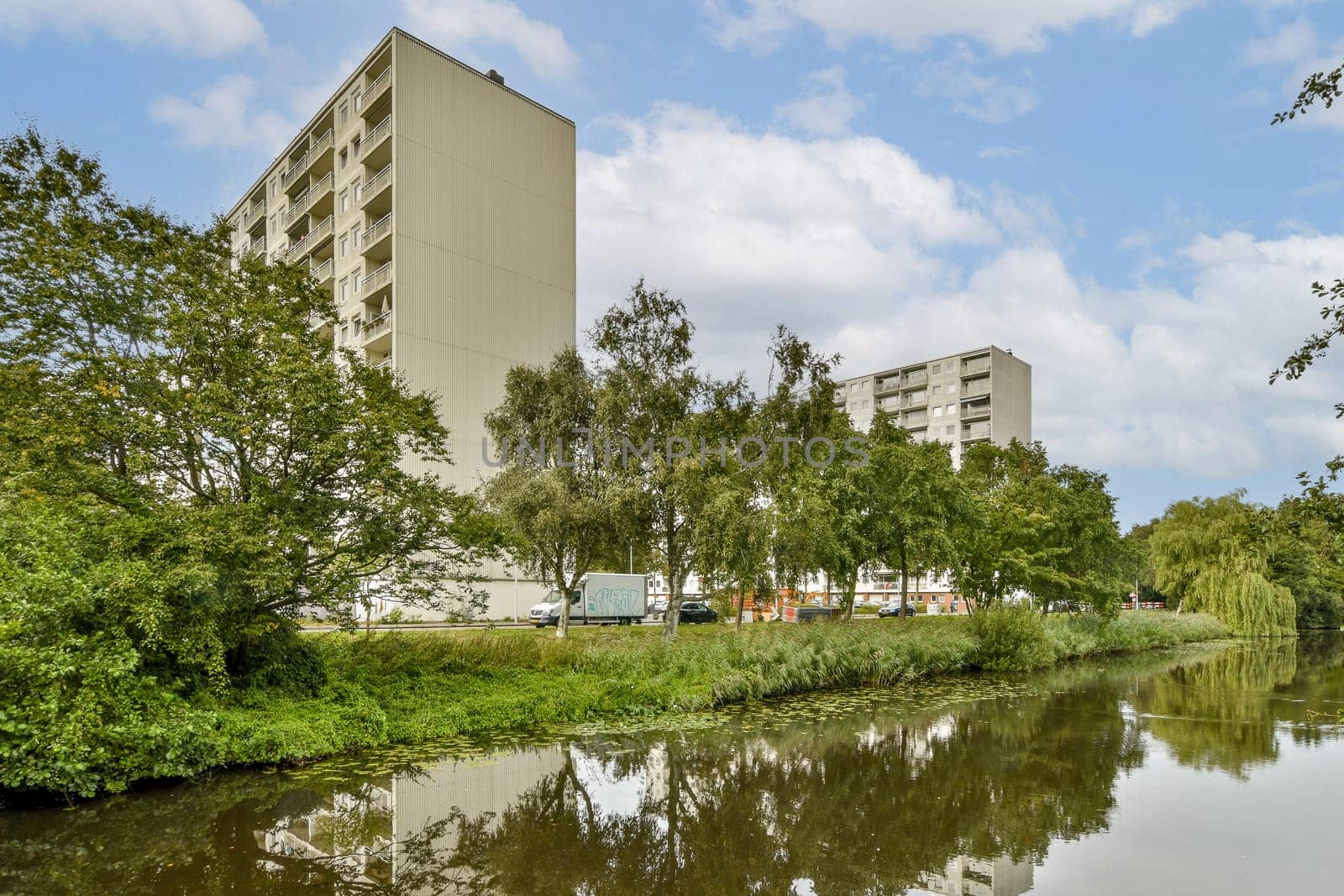 This image showcases modern buildings beside a serene river, framed by lush green trees under a cloudy sky, highlighting urban development and nature's beauty.