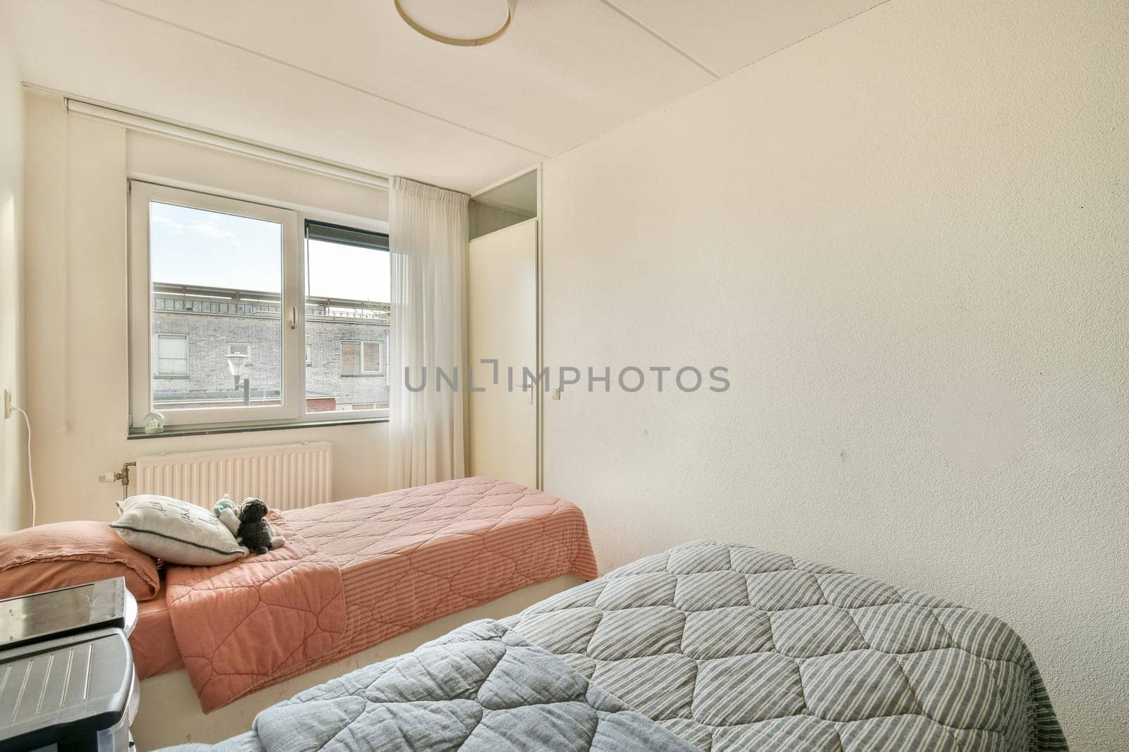 A serene bedroom featuring two neatly made beds, a large window allowing natural light, and simple, minimalistic decor that creates a calming atmosphere.