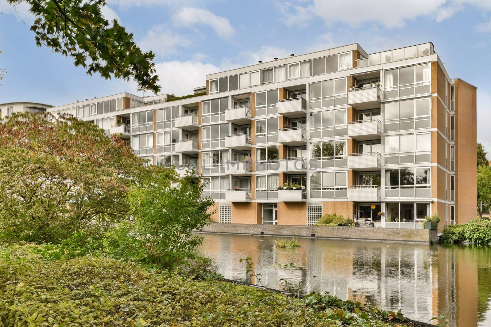 A contemporary multi-story apartment building featuring large glass windows, surrounded by lush greenery and a serene water view.