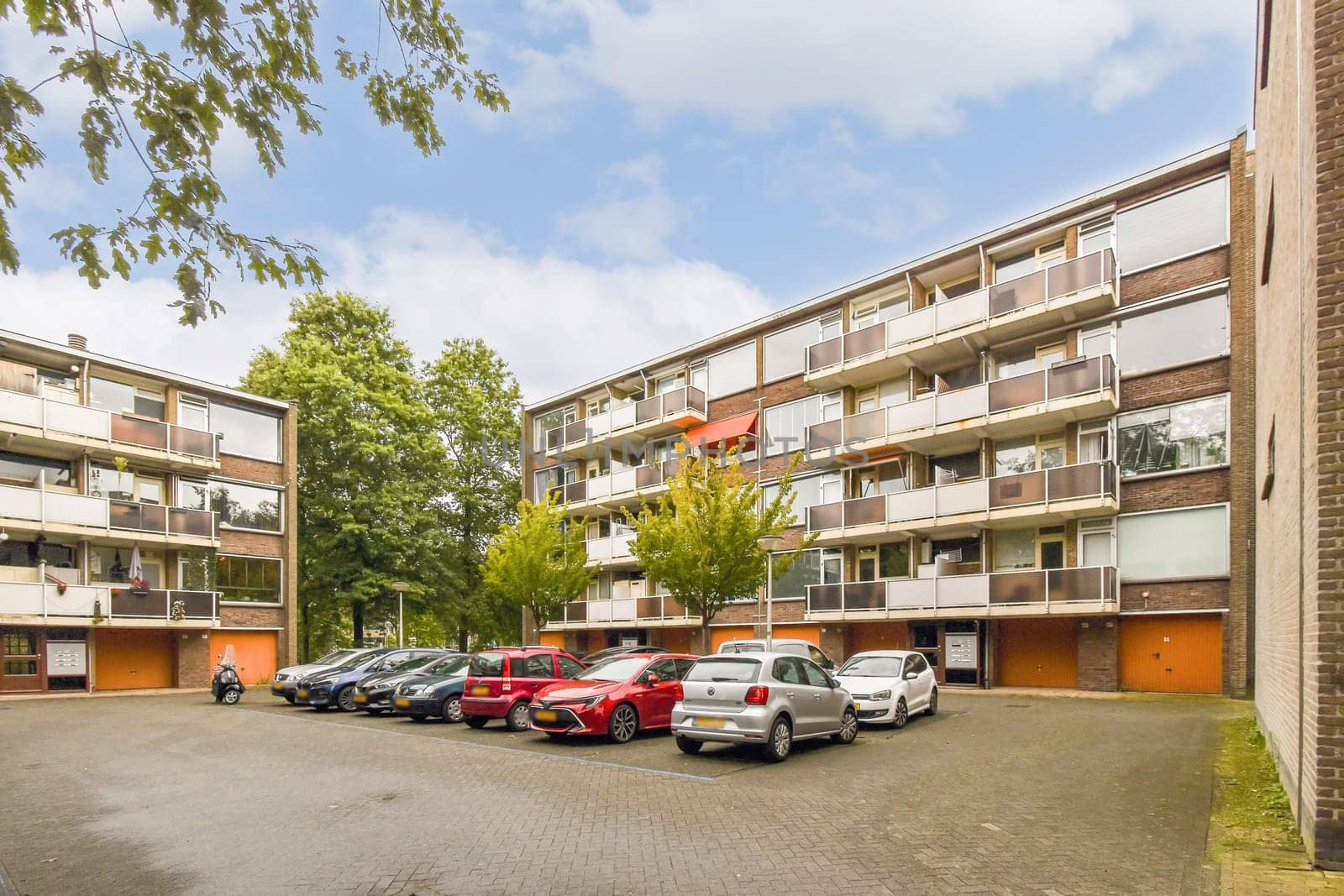 A contemporary apartment building featuring multiple floors, balconies, and an open parking area for residents. Surrounding greenery adds to its charm.