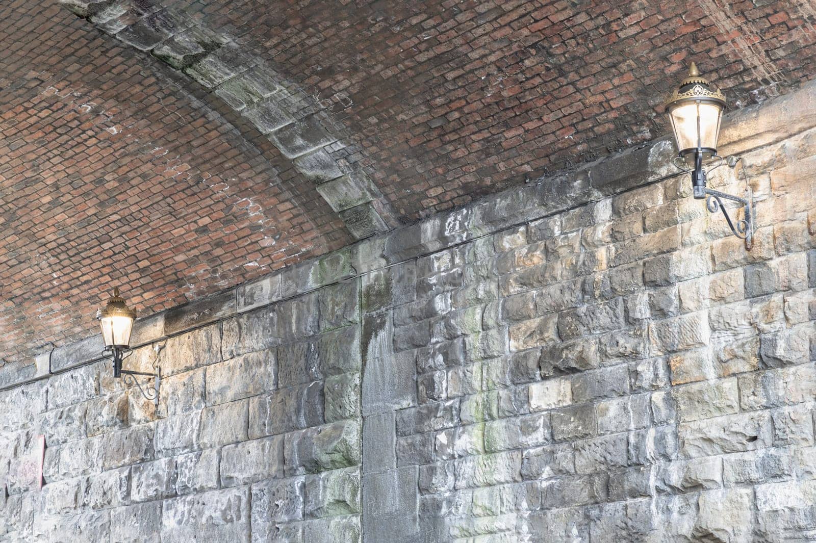 Newcastle, UK - Apr 12, 2025 - Two lantern lighting a spot on stone wall and red brick ceiling in tunnel under the high level bridge. An curved brick tunnel with lamps under the bridge, use it as your Wallpaper, Poster and Space for text, Selective focus.