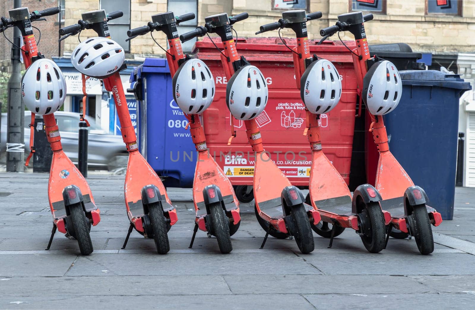 Newcastle, UK - Apr 14, 2025 - Bright orange electric scooters with white safety helmets parked on the sidewalk in downtown. Public rental e-scooter for trip and travel in center city, use it as your Wallpaper, Poster and Space for text, Selective focus.