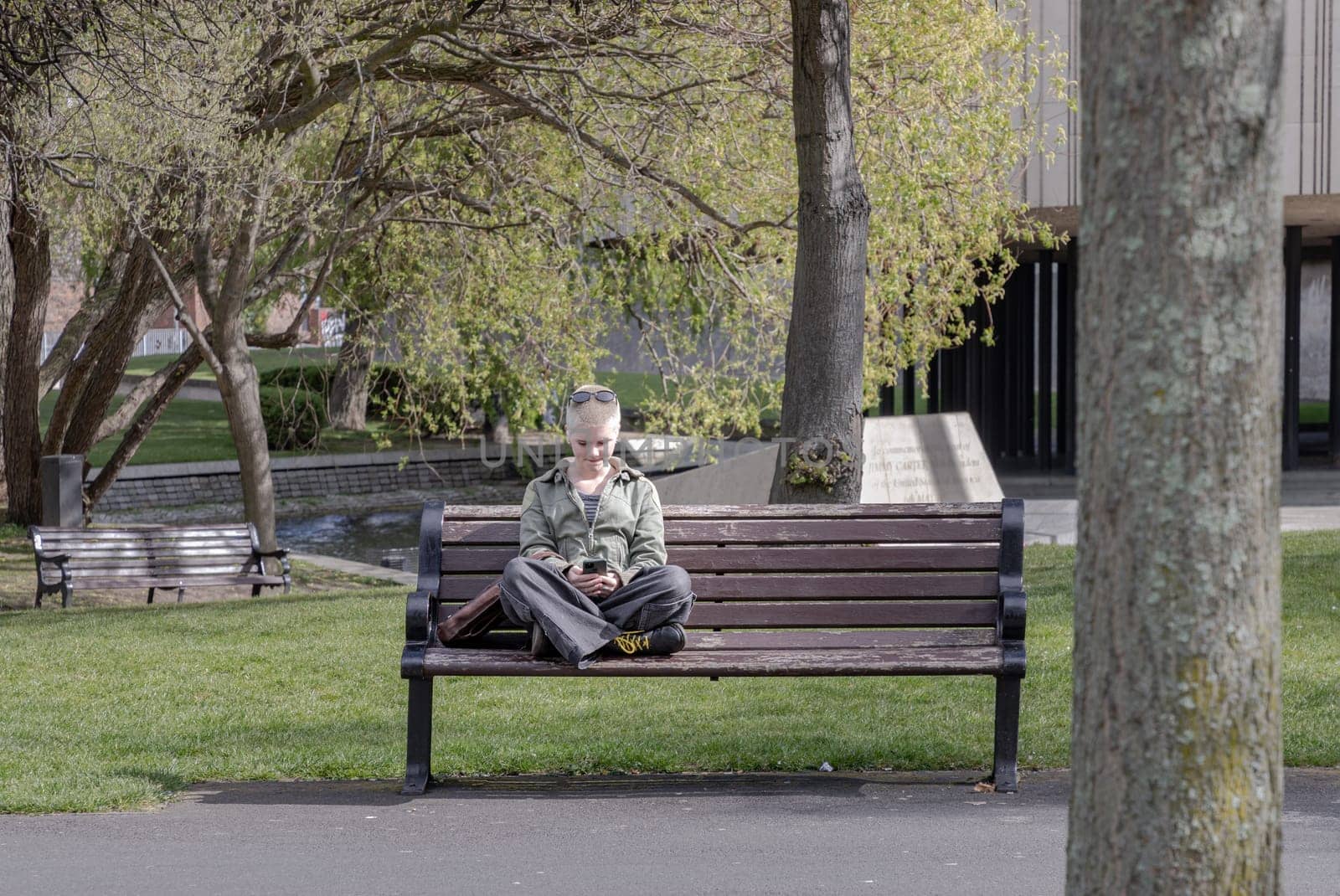 Newcastle, UK - Apr 14, 2025 - A young woman blonde short hair wearing jacket and jeans top with sunglasses on her head is sits on a old bench in a quiet park during a warm afternoon. looking down at her smartphone, use it as your Wallpaper, Poster and Space for text, Selective focus.
