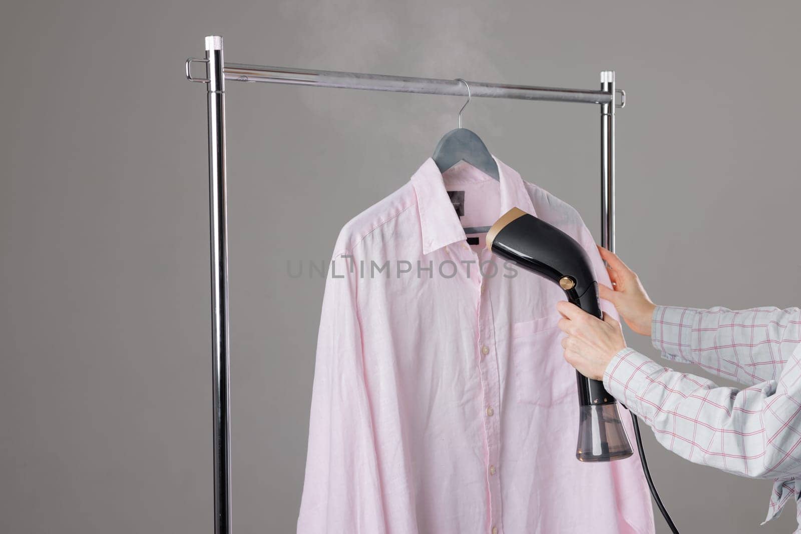 Woman steaming pink shirt on hanger using handheld garment steamer to remove wrinkles, home laundry care.