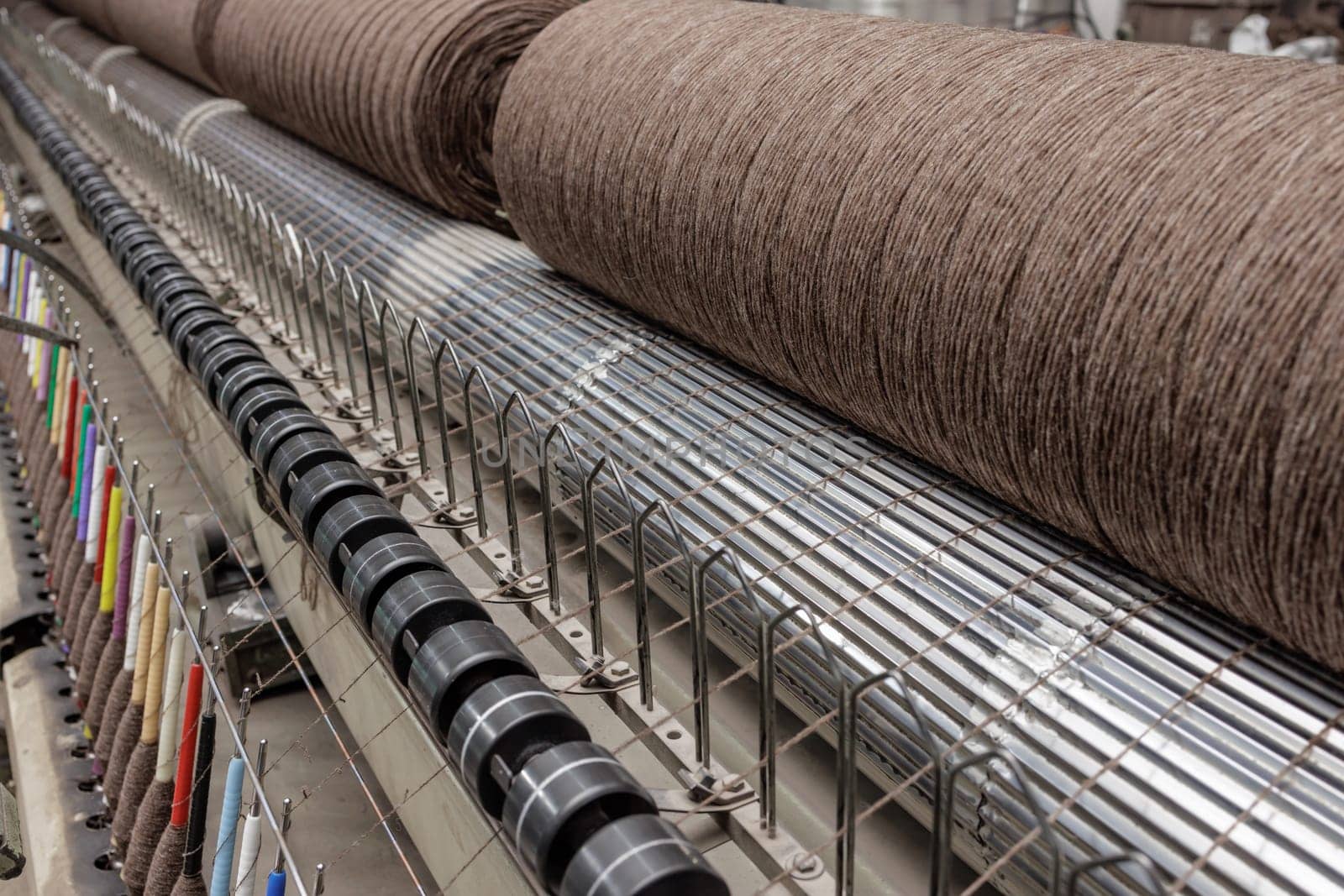 Industrial textile machine with large brown yarn spool in motion, producing fabric with colorful thread guides.