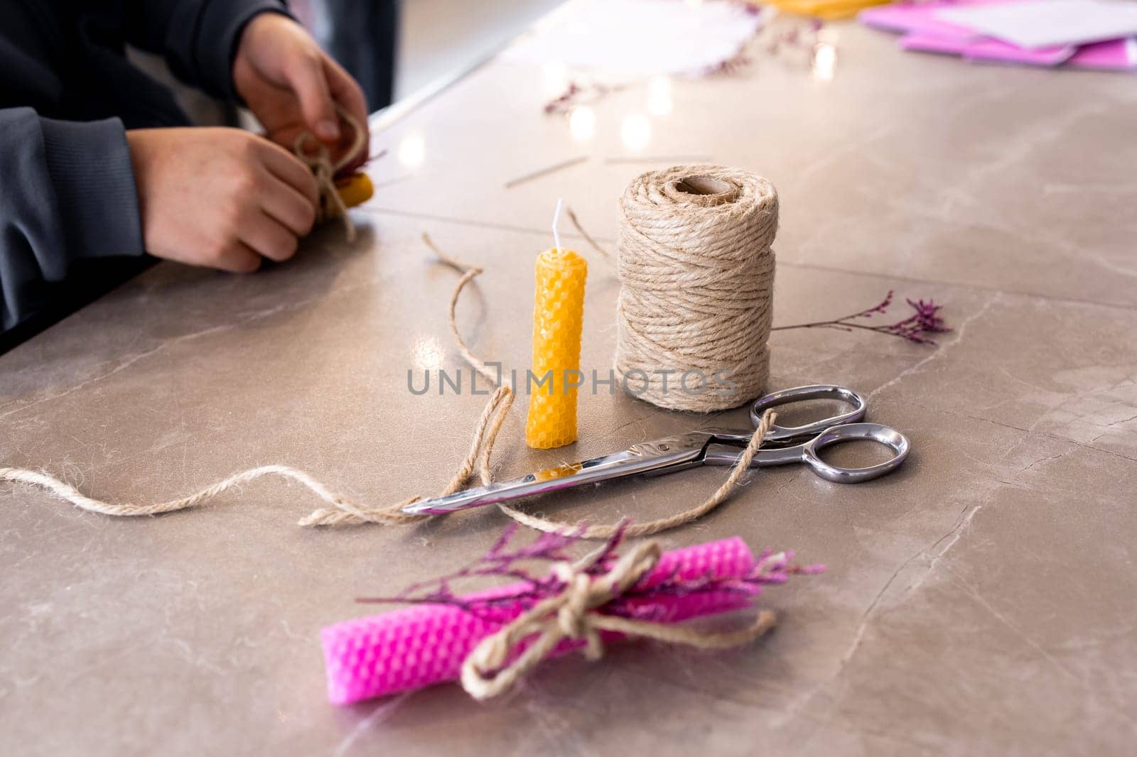 Young hands crafting beeswax candle with natural twine on marble surface.
