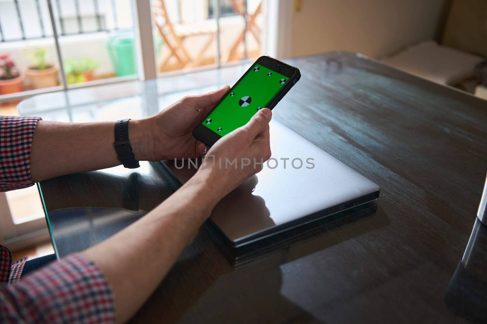Man interacts with a smartphone displaying a green screen, seated at a table indoors. Nearby, a sleek laptop rests on the desk, depicting a workspace setting. Natural light fills the room.