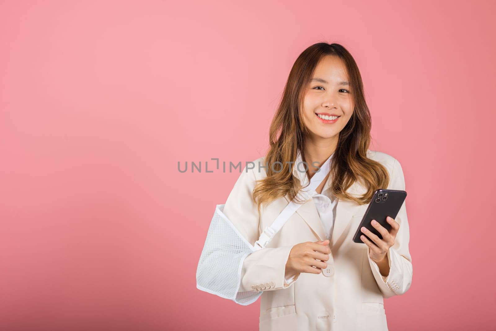 Woman confident smiling broken arm after accident and wear arm splint for treatment and hold smartphone, happy Asian female sling supported hand isolated on pink background, social media, copy space