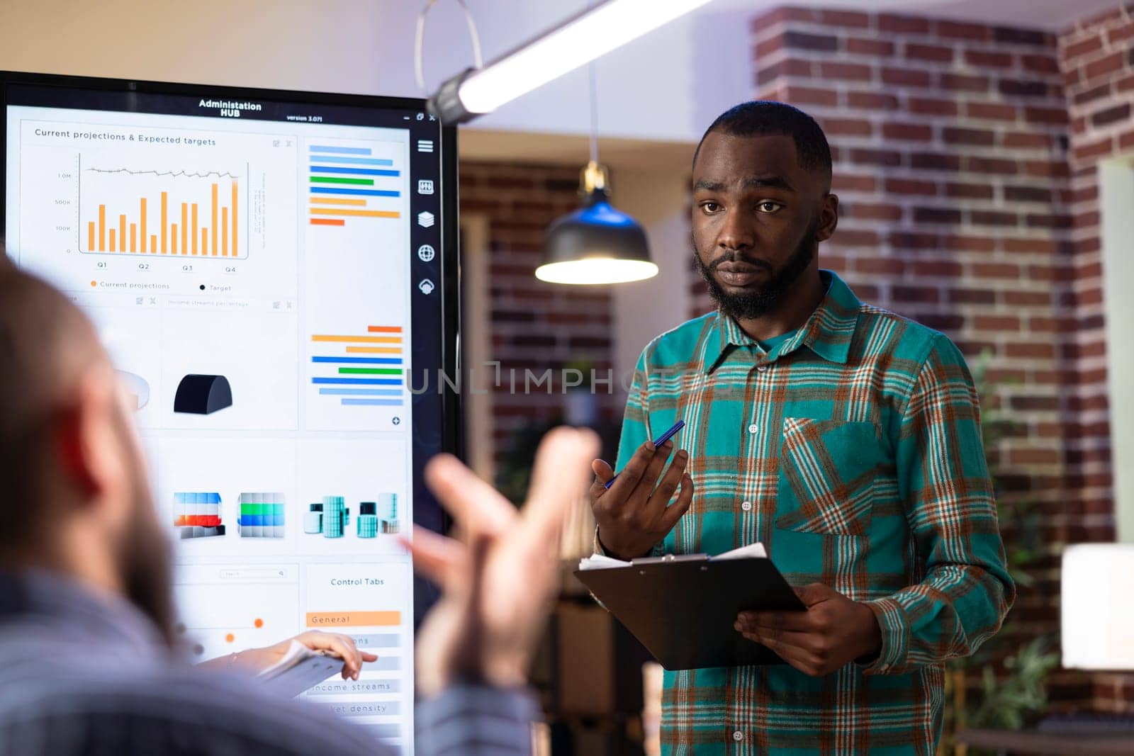 Black male entrepreneur presents strategy ideas on digital screen, leading a brainstorming session in brick wall office. African american businessman engaging his team in collaborative meeting.